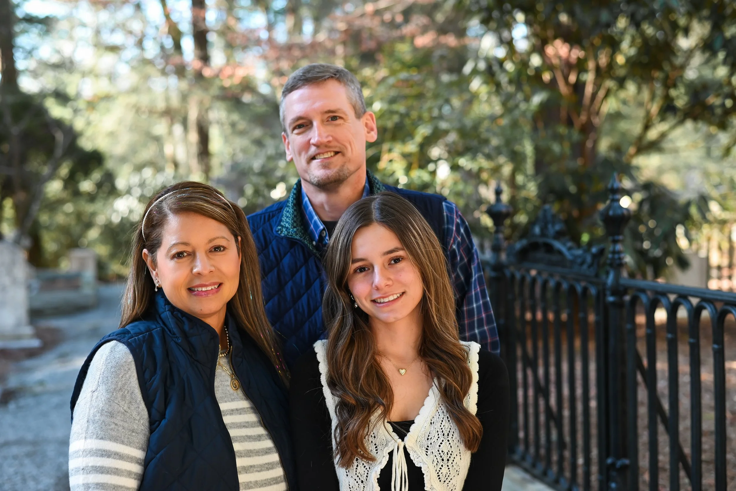 A family of three, including a woman, a man, and a teenage girl, standing outdoors in a park-like setting with trees and a decorative iron fence in the background during fall.
