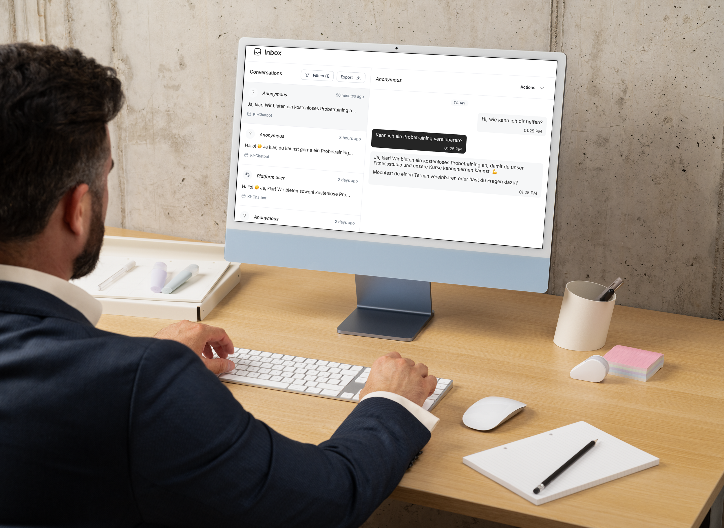 Ein Mann arbeitet an einem Schreibtisch mit einem Computer, der einen Chat-Brom darstellt. Auf dem Tisch befinden sich Notizblock, Stifte, Post-it-Blöcke, eine Maus, und eine Kerze im Becher.