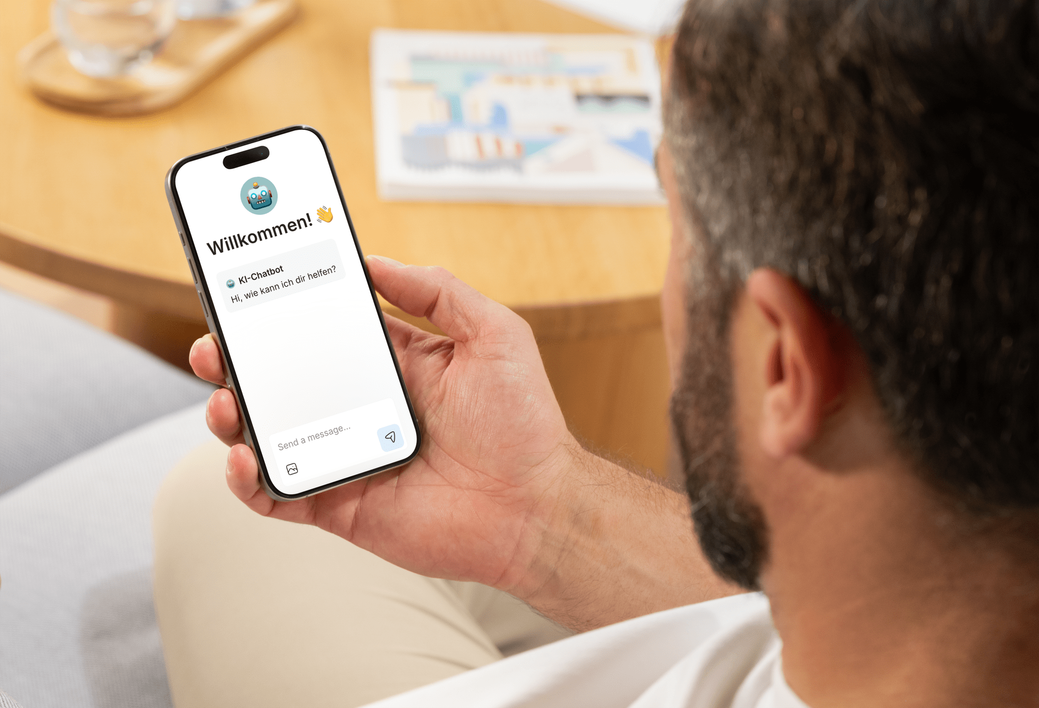Ein Mann hält ein Smartphone mit einer WhatsApp-Chat-Nachricht, in der 'Willkommen! 👋' und ein Roboter-Emoji sowie die Frage 'Hi, wie kann ich dir helfen?' zu sehen sind, auf einem Holztisch mit Zeitschriften im Hintergrund.