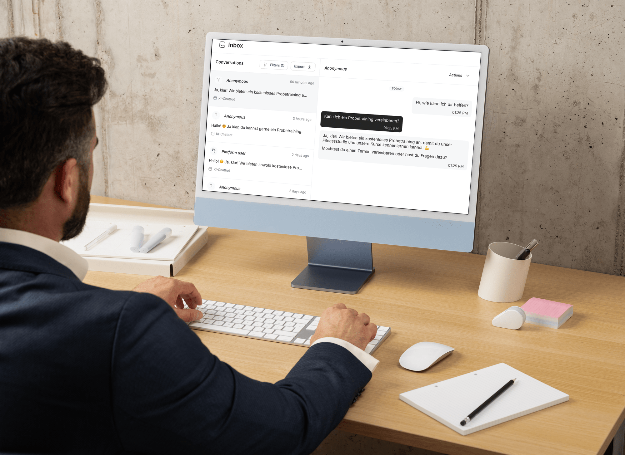 Ein Mann im Anzug arbeitet an einem Computer in einem modernen Büro. Der Bildschirm zeigt eine Chat-Konversation auf Deutsch.