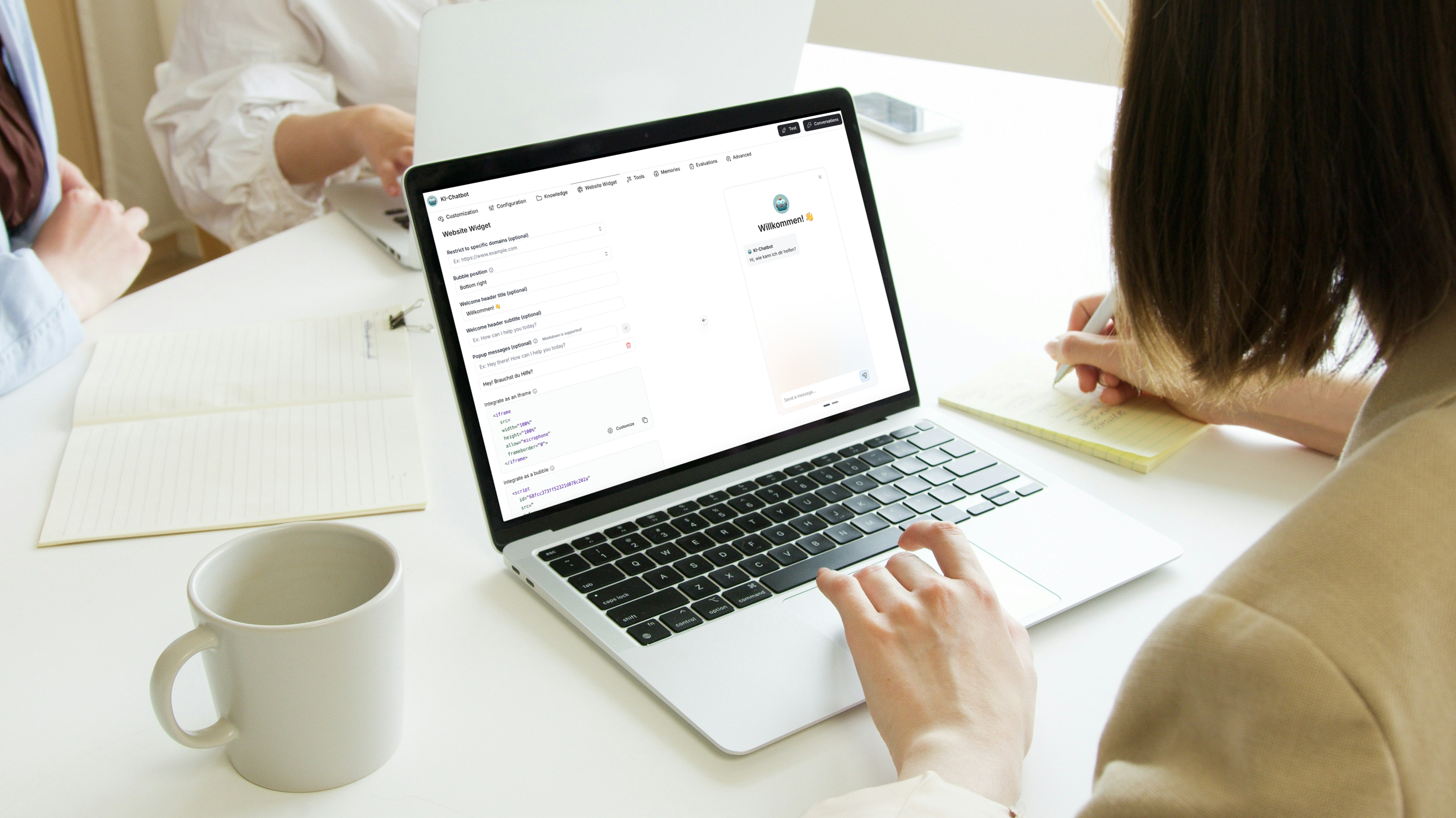 Person tippt auf einem Laptop mit einem Chatfenster und einer Willkommensnachricht, während andere Personen am Tisch Notizen machen, eine Tasse und Notizen sichtbar.