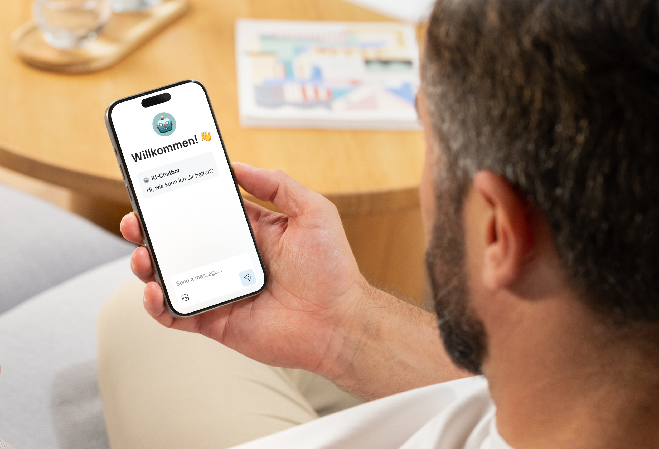 Ein Mann hält ein Smartphone mit einem Chatbot-Dialog, auf dem steht 'Willkommen! Wie kann ich dir helfen?'. Im Hintergrund ein Tisch mit einem Glas Wasser und einem Magazin.