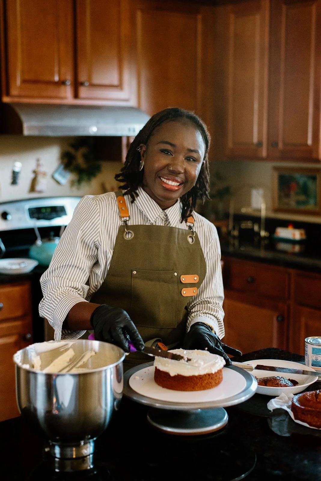 A woman smiling and wearing an apron and gloves, decorating a cake in a kitchen.
