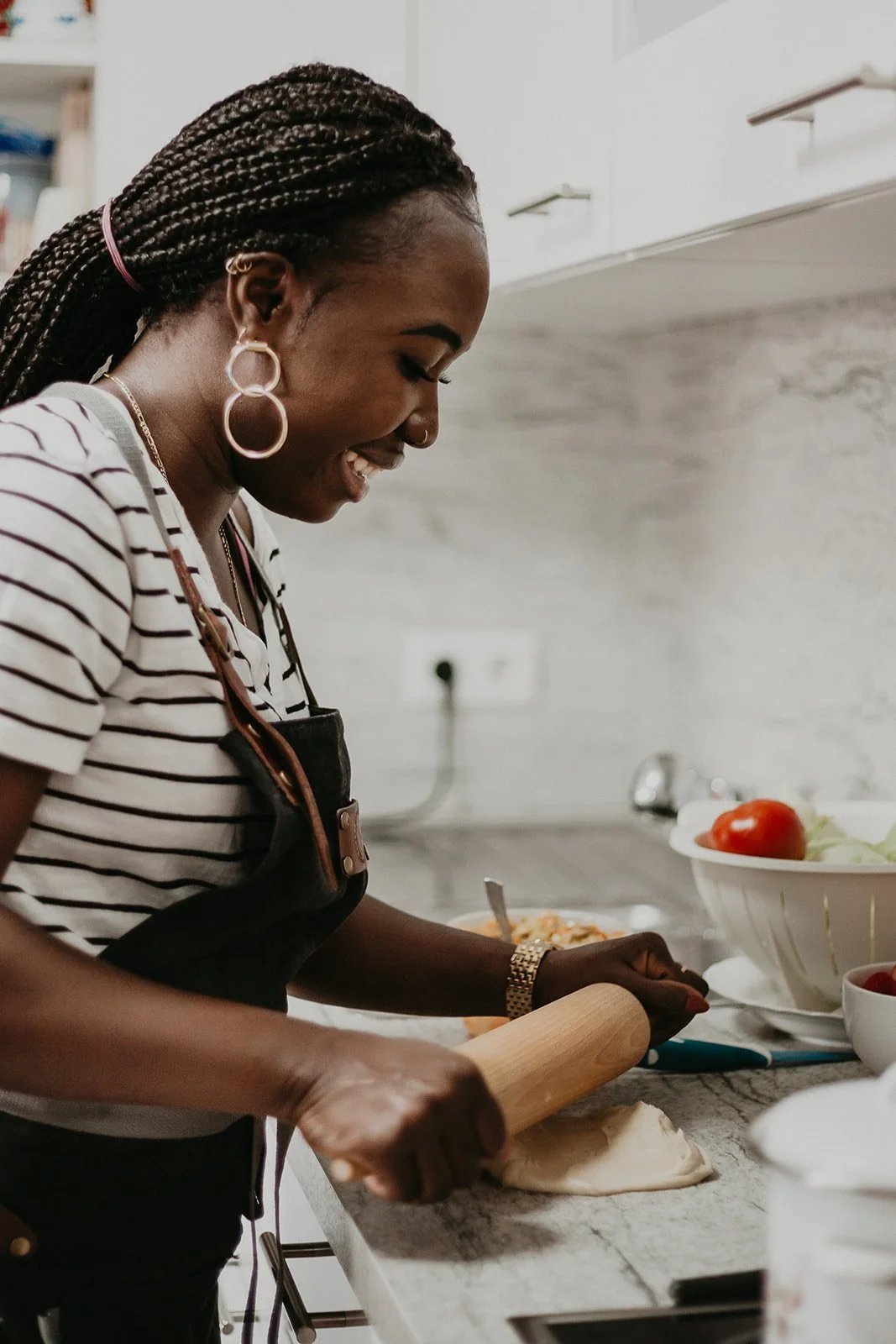 A woman with braided hair and large hoop earrings rolling dough on a kitchen countertop, smiling while preparing food.