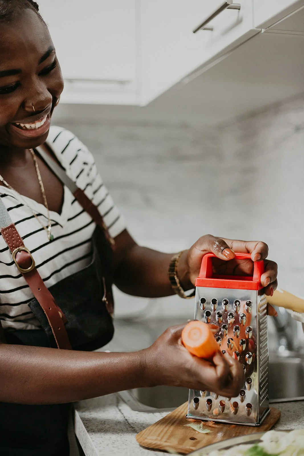 A woman with dark skin and a broad smile, wearing a striped shirt and an apron, grates a carrot using a box grater in a kitchen with white cabinets.