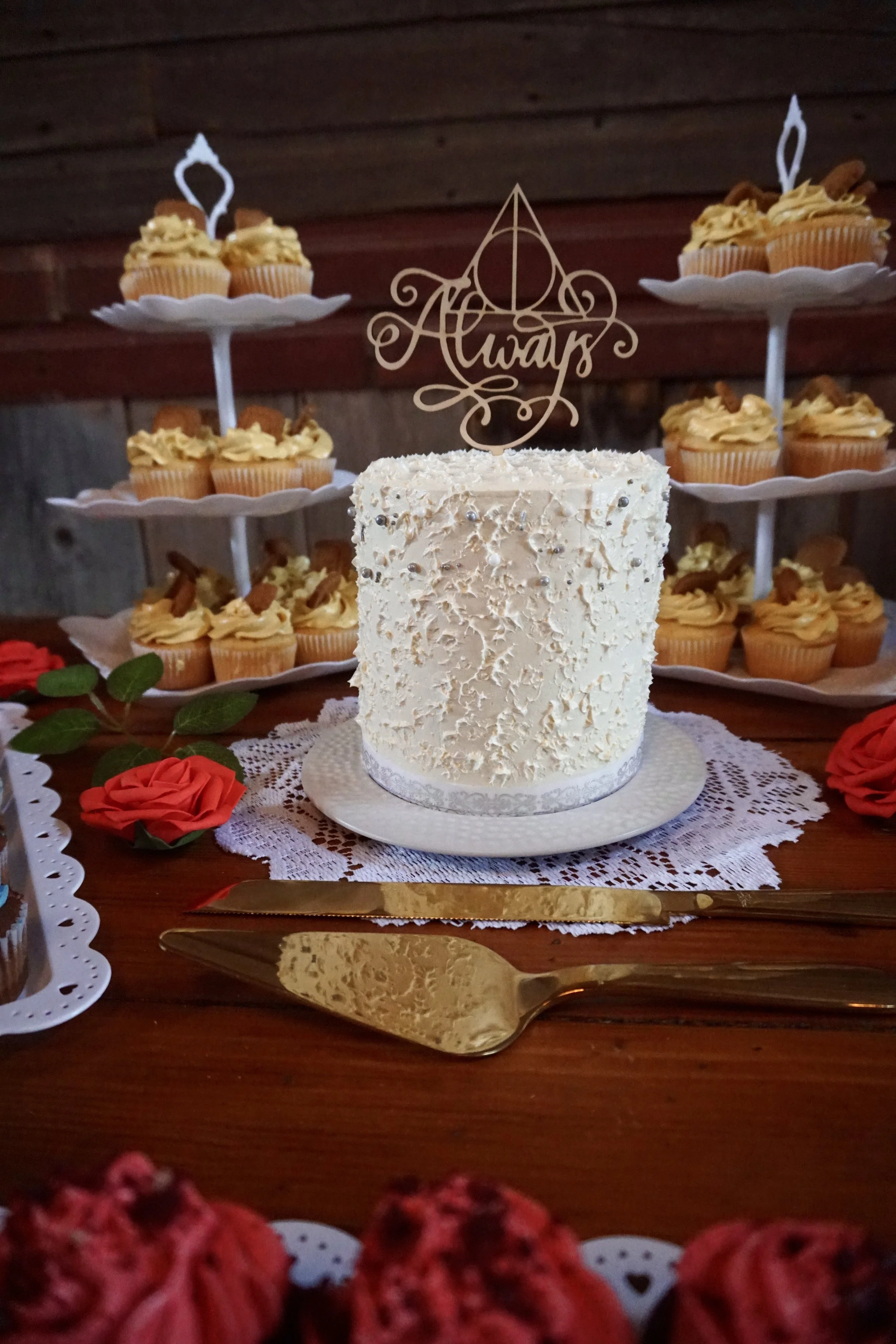 A white frosted cake with silver embellishments and a topper that says 'Awards' on a white doily, with cupcakes on a stand and decorative roses around it.