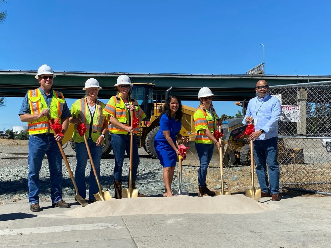 Martha Guerrero and people at a groundbreaking ceremony with shovels, some wearing safety vests and helmets, standing in front of construction equipment and a bridge.