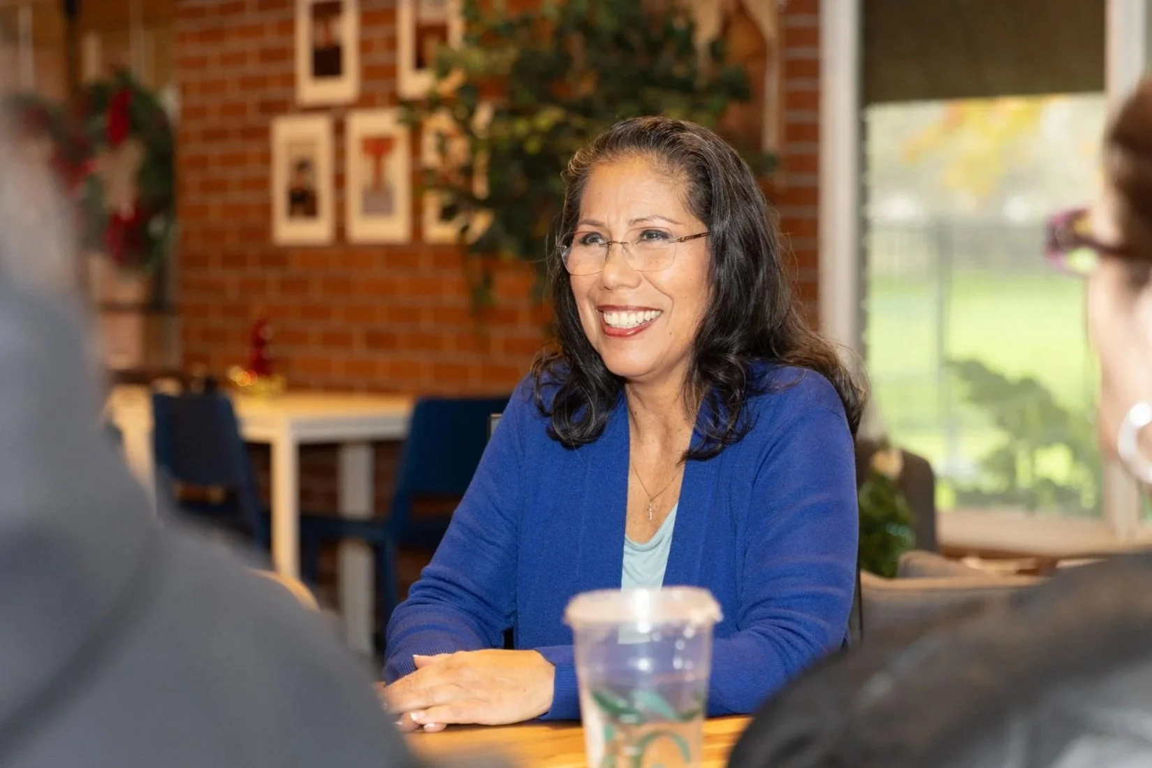 Martha Guerrero with long dark hair, glasses, and a blue blazer smiling while sitting at a table in a cozy room with brick walls, decorative framed pictures, and a large window showing greenery outside.