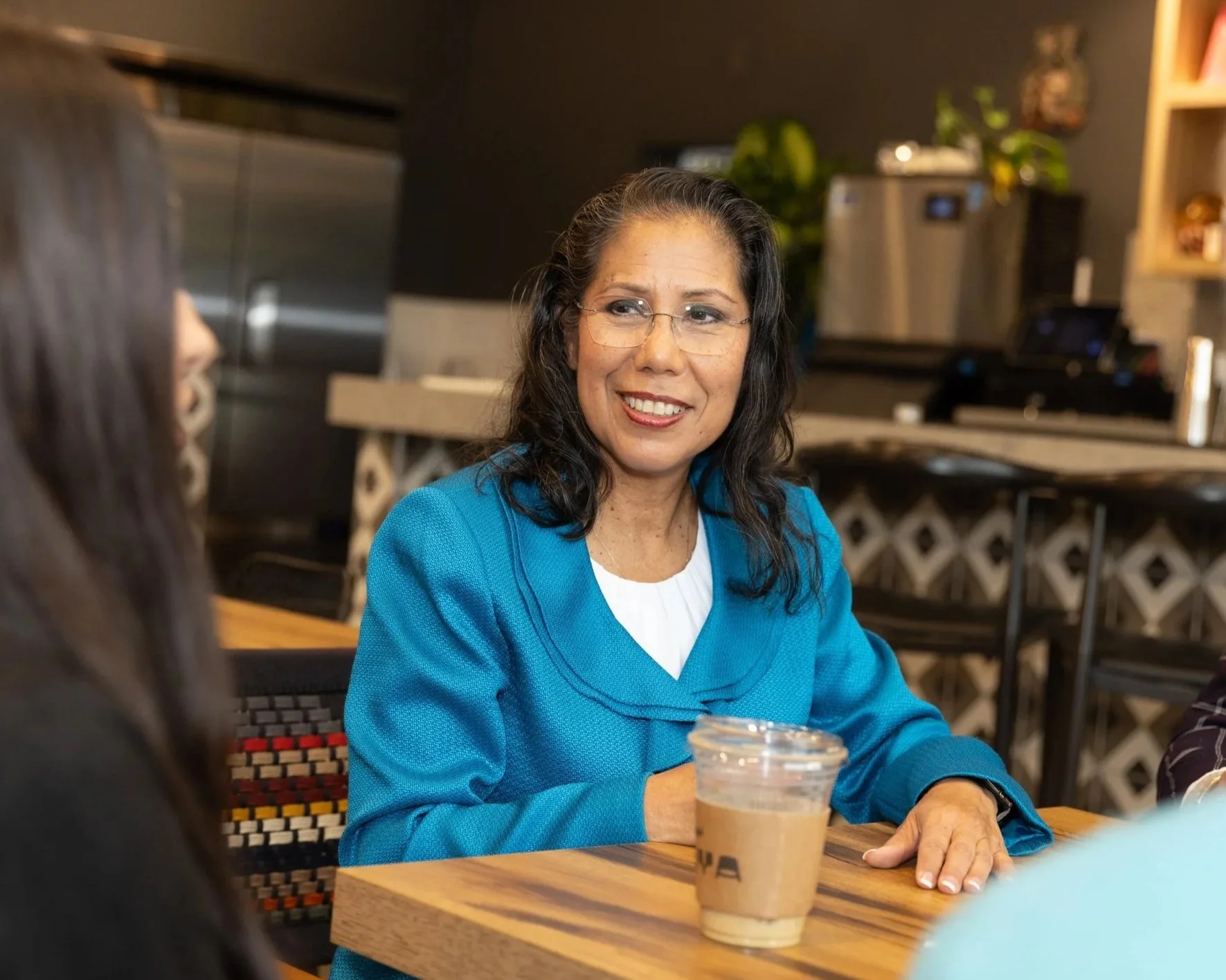 Martha Guerrero in a blue blazer sitting at a wooden table with a coffee cup, smiling and talking to someone.