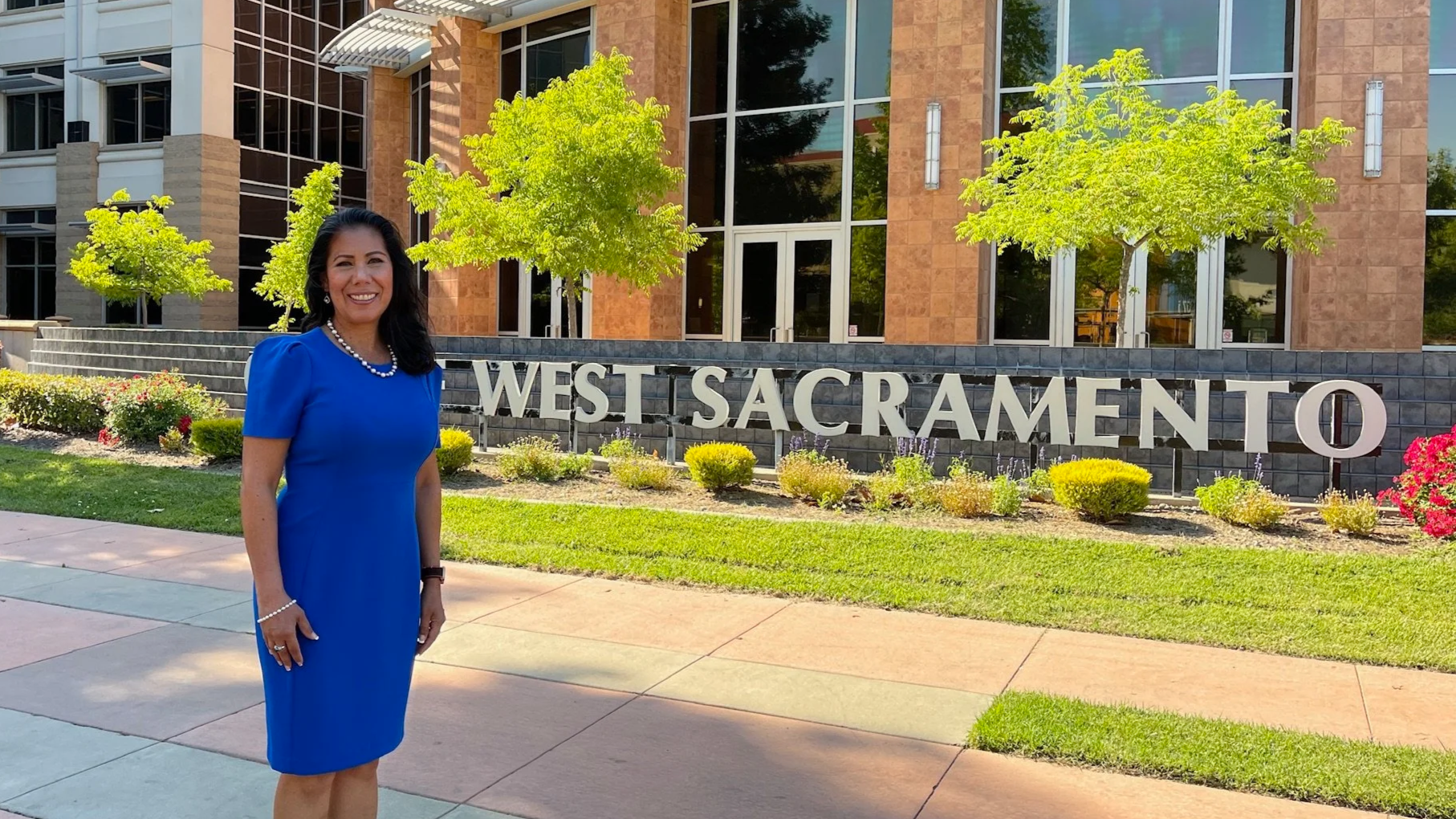 Martha Guerrero in a blue dress standing in front of the West Sacramento sign outside a modern building with glass windows and trees.