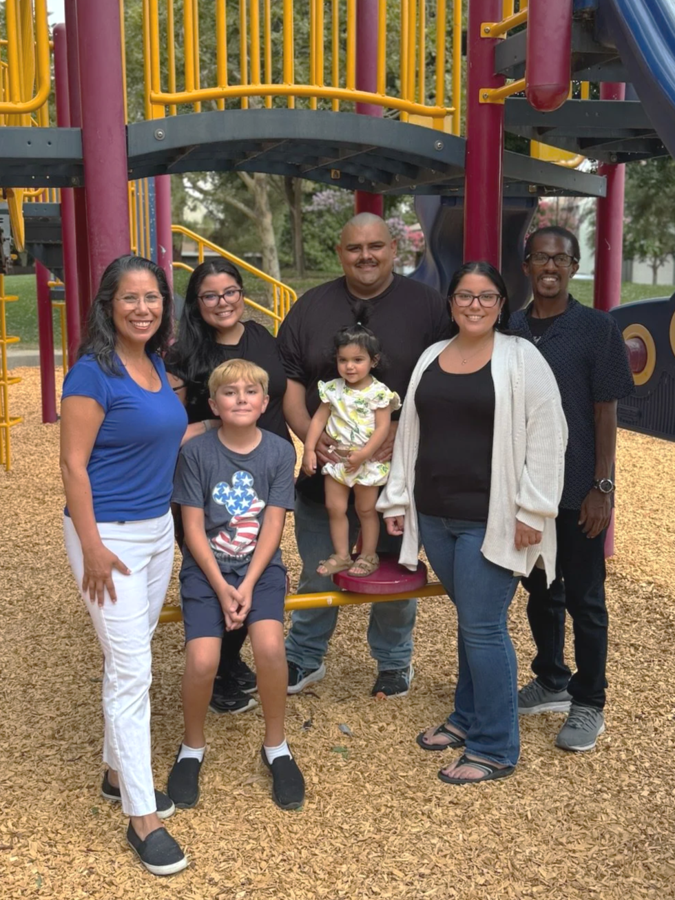 Group of seven people, including children and adults and Mayor Martha Guerrero, standing on playground equipment with slides and climbing structures, outdoors on a wood-chip surface.