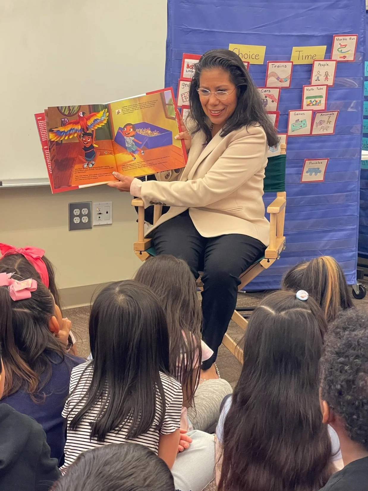 Martha Guerrero reads a colorful children's book to a group of children sitting on the floor in a classroom. Martha Guerrero is smiling and holding the book open, showing illustrations of children and a parrot.
