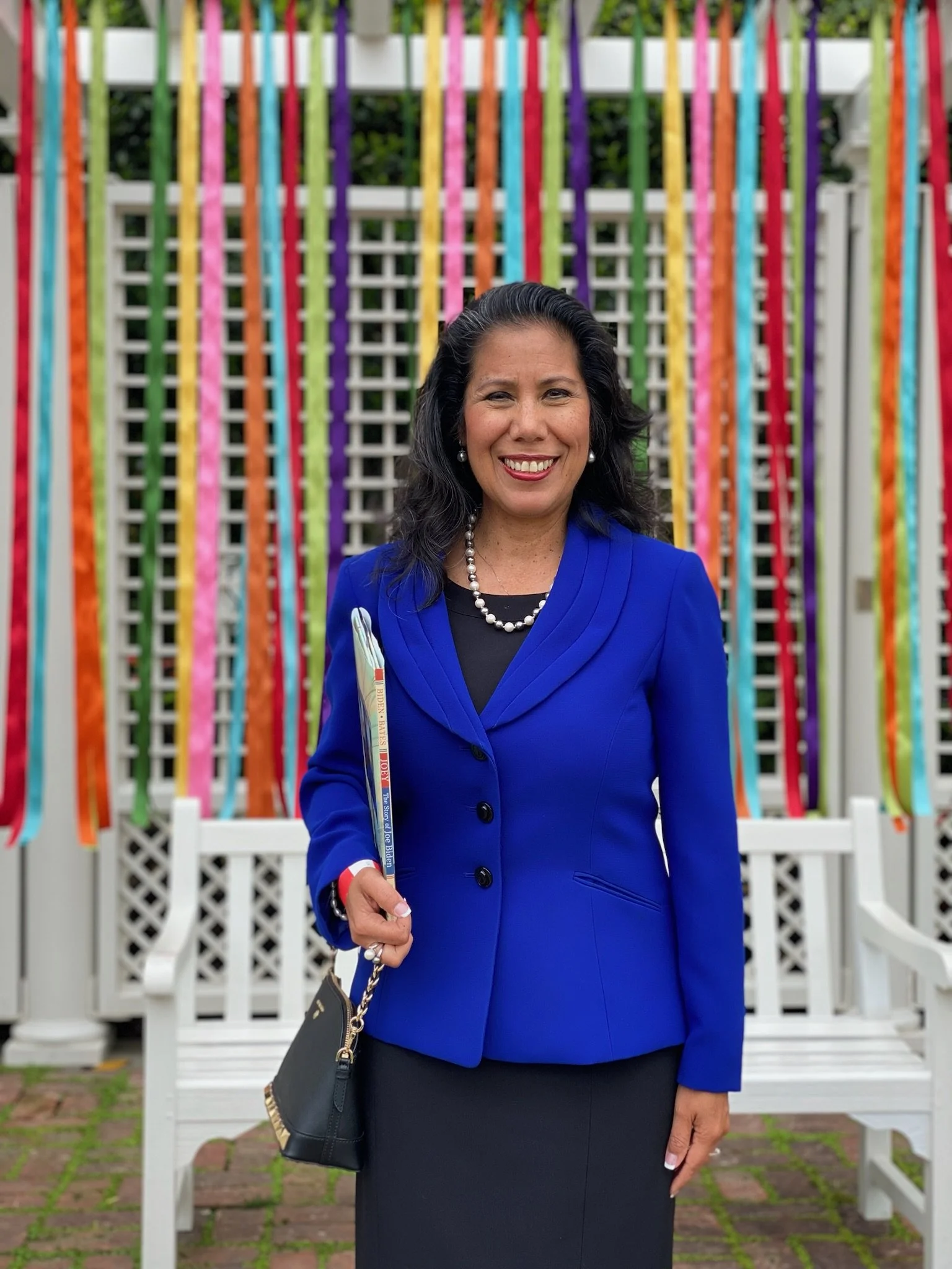 Martha Guerrero wearing a royal blue blazer, black skirt, pearl necklace and earrings, holding a small purse and a rectangular object, standing in front of colorful streamers and white outdoor furniture.