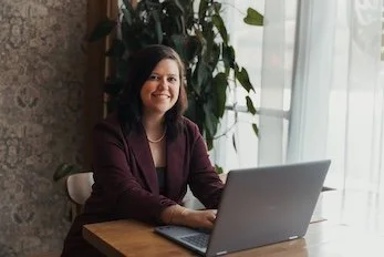 Woman sitting at a desk with a laptop in a well-lit room, smiling at the camera.