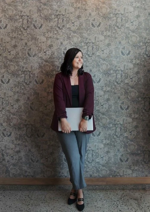 Woman standing with a laptop in front of a patterned wall, wearing a maroon blazer, black top, gray pants, and black shoes.