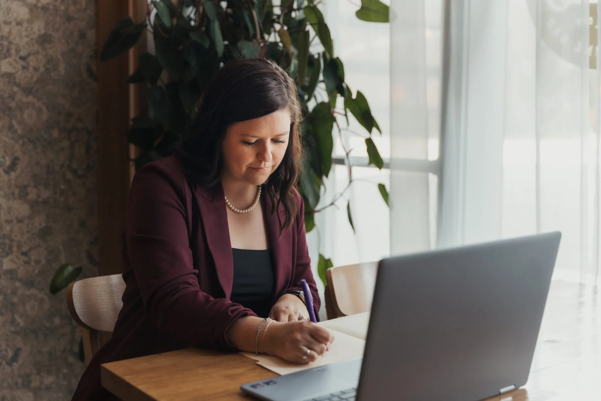 Woman in burgundy blazer writing in notebook at a wooden table with laptop nearby.