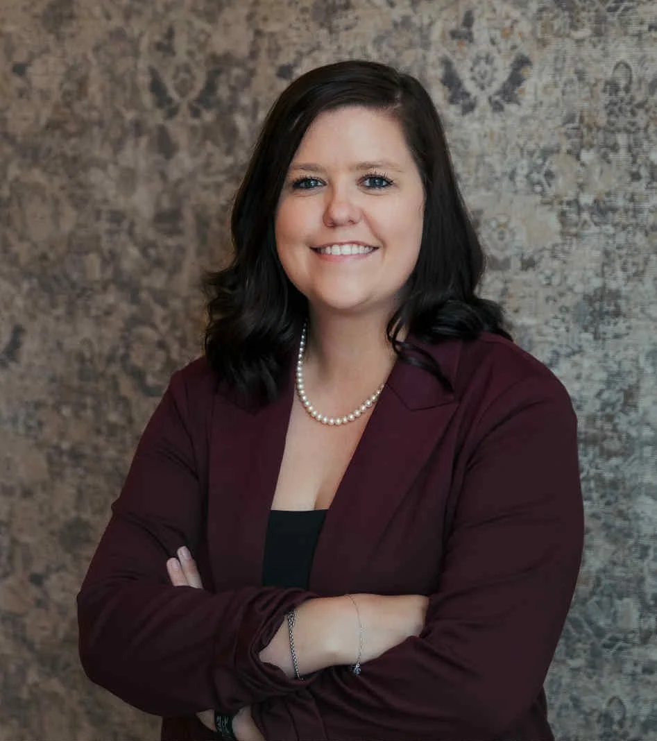 A woman with shoulder-length dark hair, wearing a maroon blazer, pearl necklace, and a black top, standing with arms crossed in front of a textured wall, and smiling at the camera.