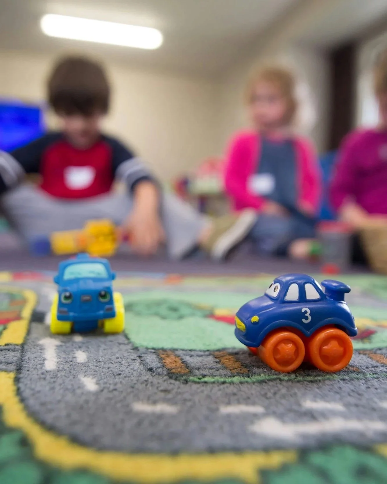 Close-up of two small toy cars on a colorful playmat with children blurred in the background.