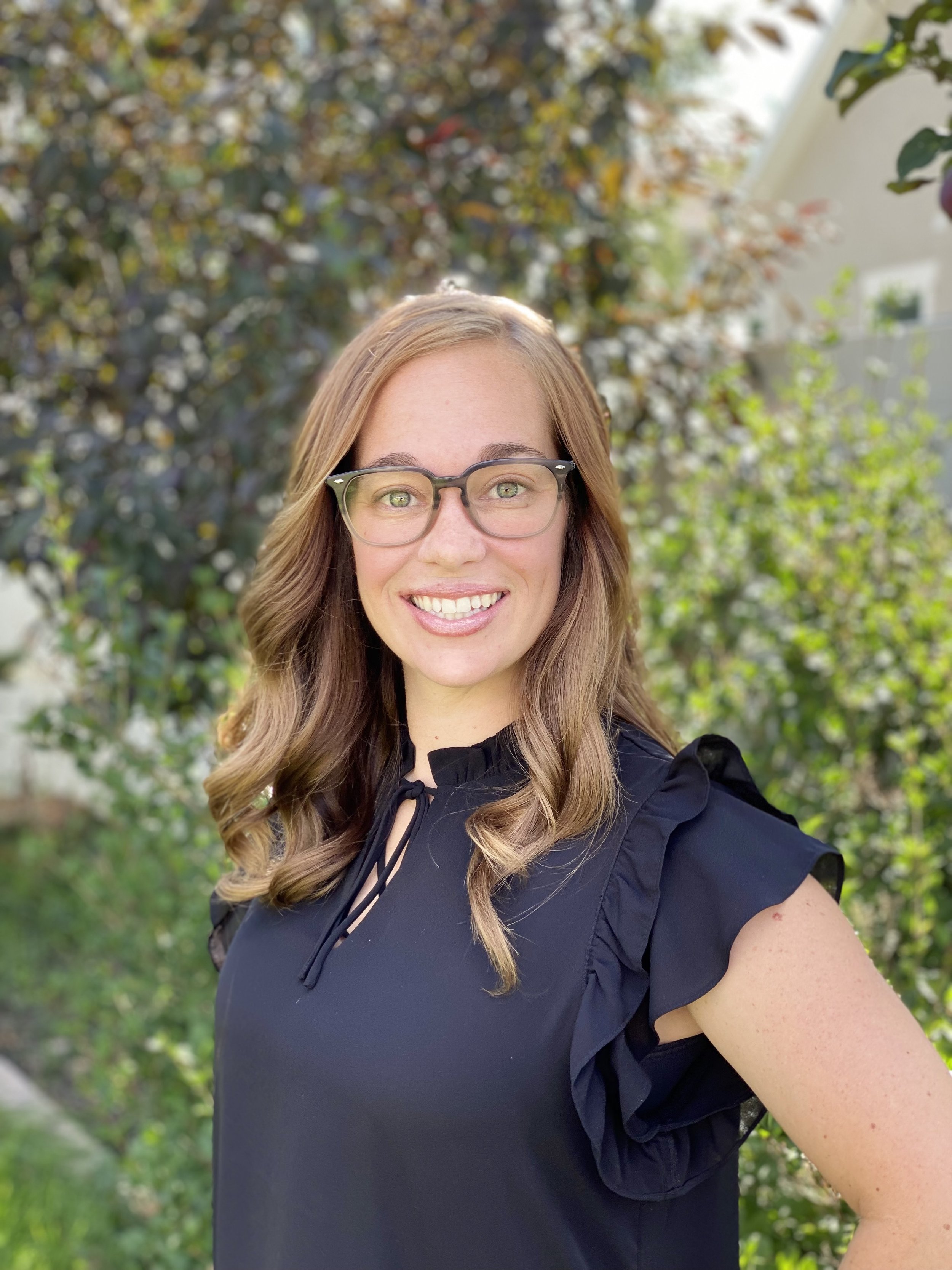 A woman with light brown, wavy hair, wearing glasses and a black ruffled blouse, smiling outdoors on a sunny day with green foliage and a white building in the background.