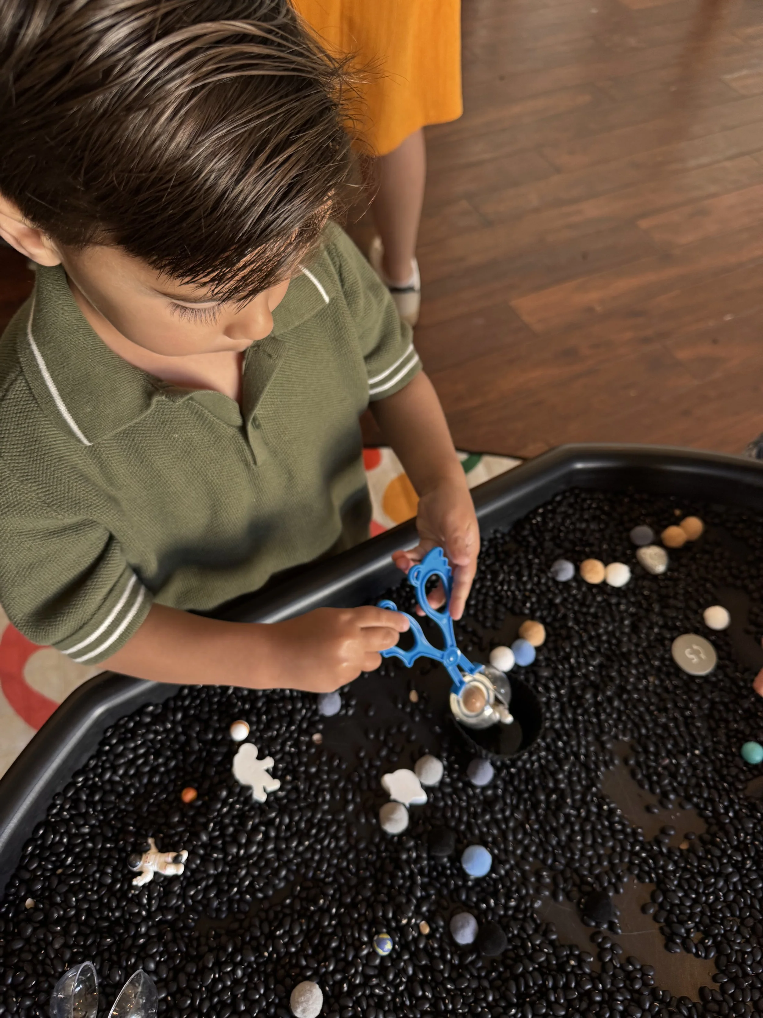 Child playing with a sensory table  filled with  black bean's and space items in a room with hardwood flooring.