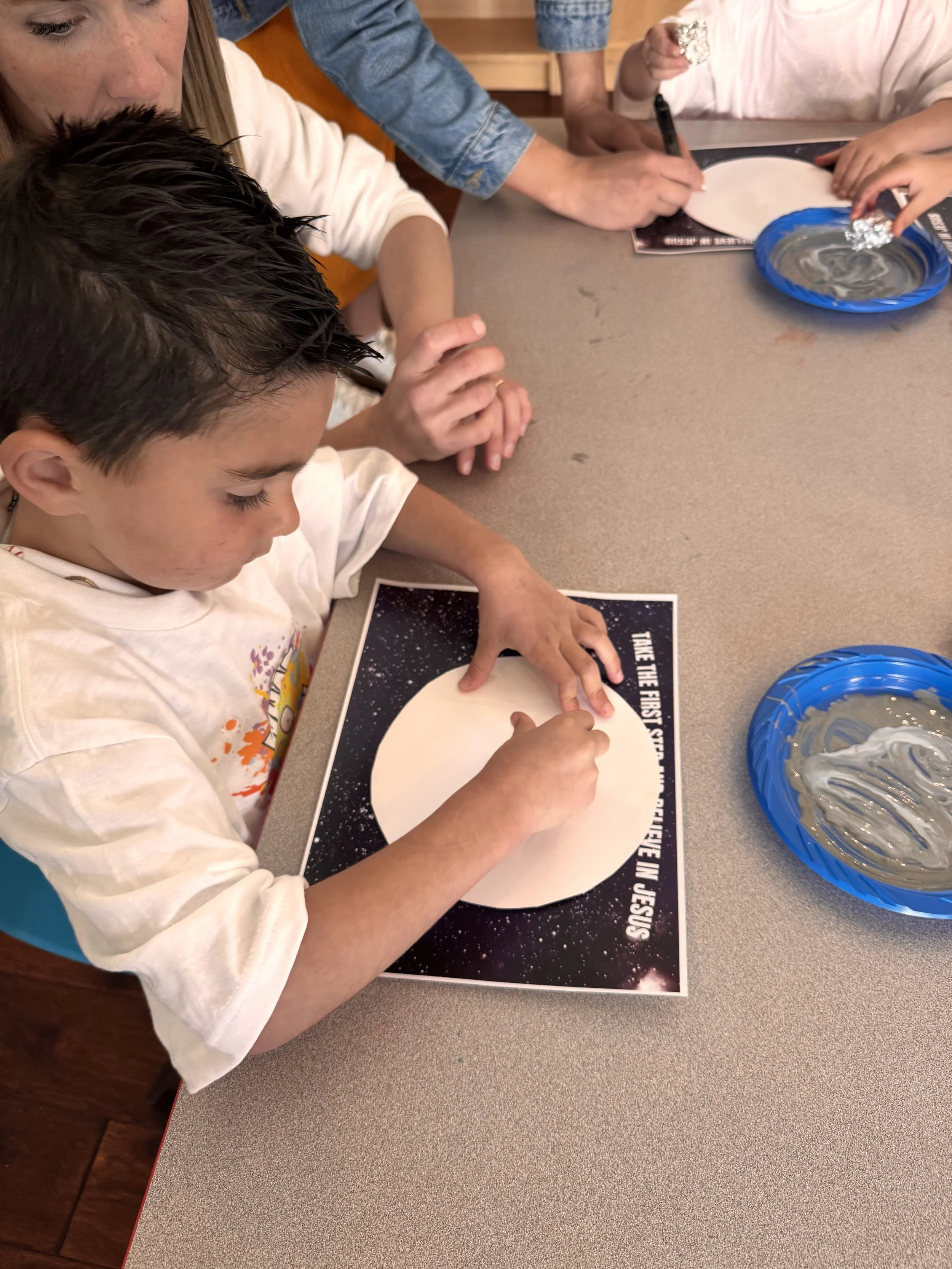 Children and adult creating a space-themed craft together at a table, with one child drawing on a paper circle, and a bowl of silver paint nearby.