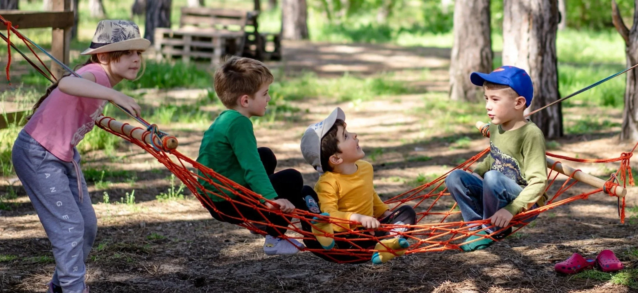 Four children playing on a hammock in a wooded outdoor area