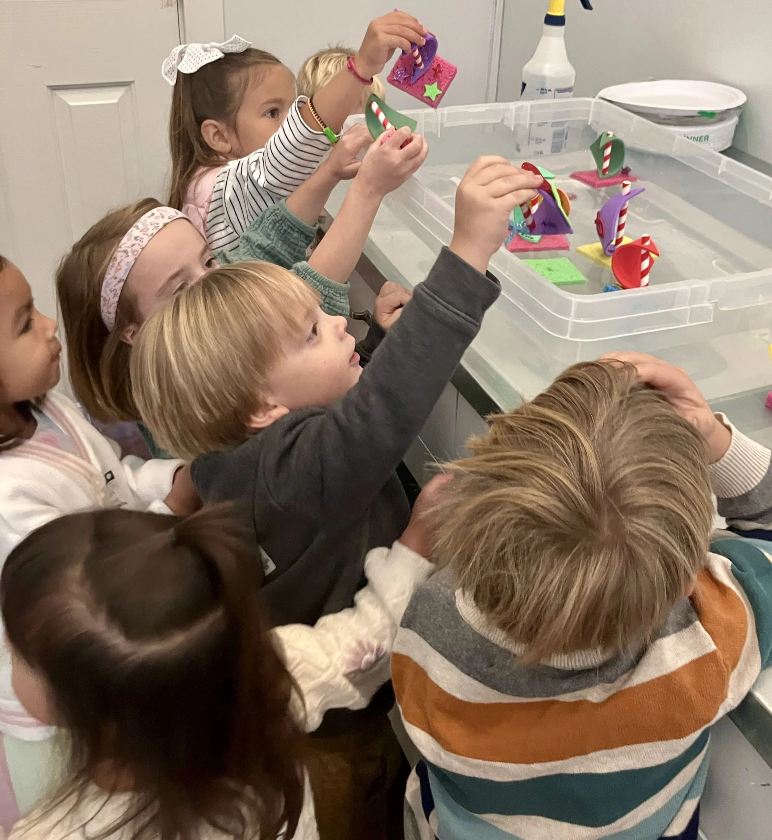 A group of children reaching into a clear plastic container filled with colorful foam crafts, with a white door and a cleaning spray bottle in the background.