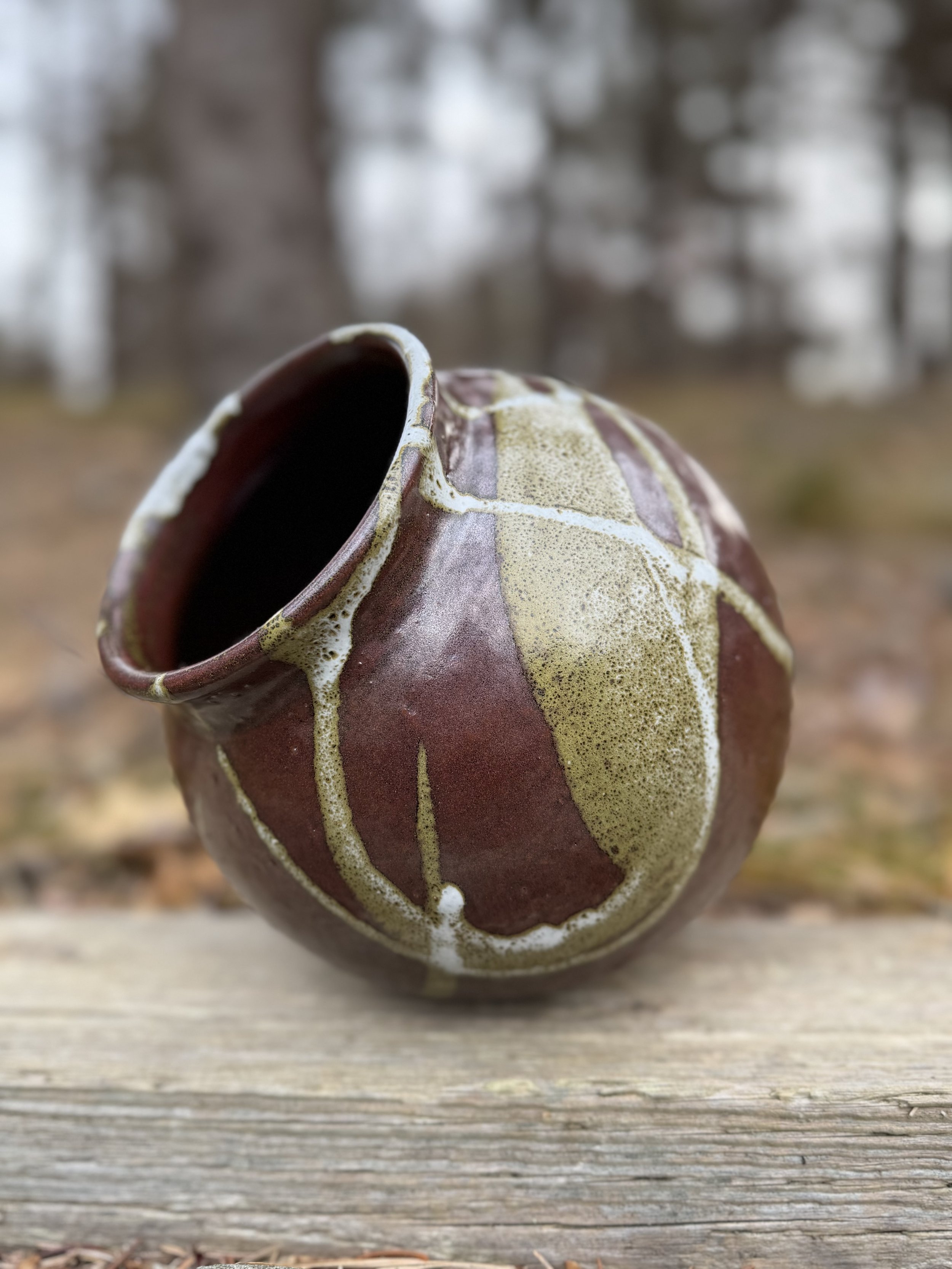 A ceramic vase with a geometric pattern in maroon and beige, filled with a dark liquid, placed on a rustic wooden surface outdoors with a blurred natural background.