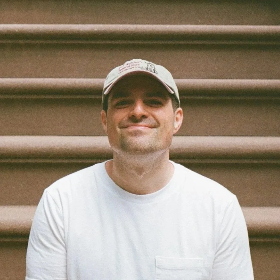 A man smiling and wearing a baseball cap and white t-shirt, sitting on brown stadium seats.