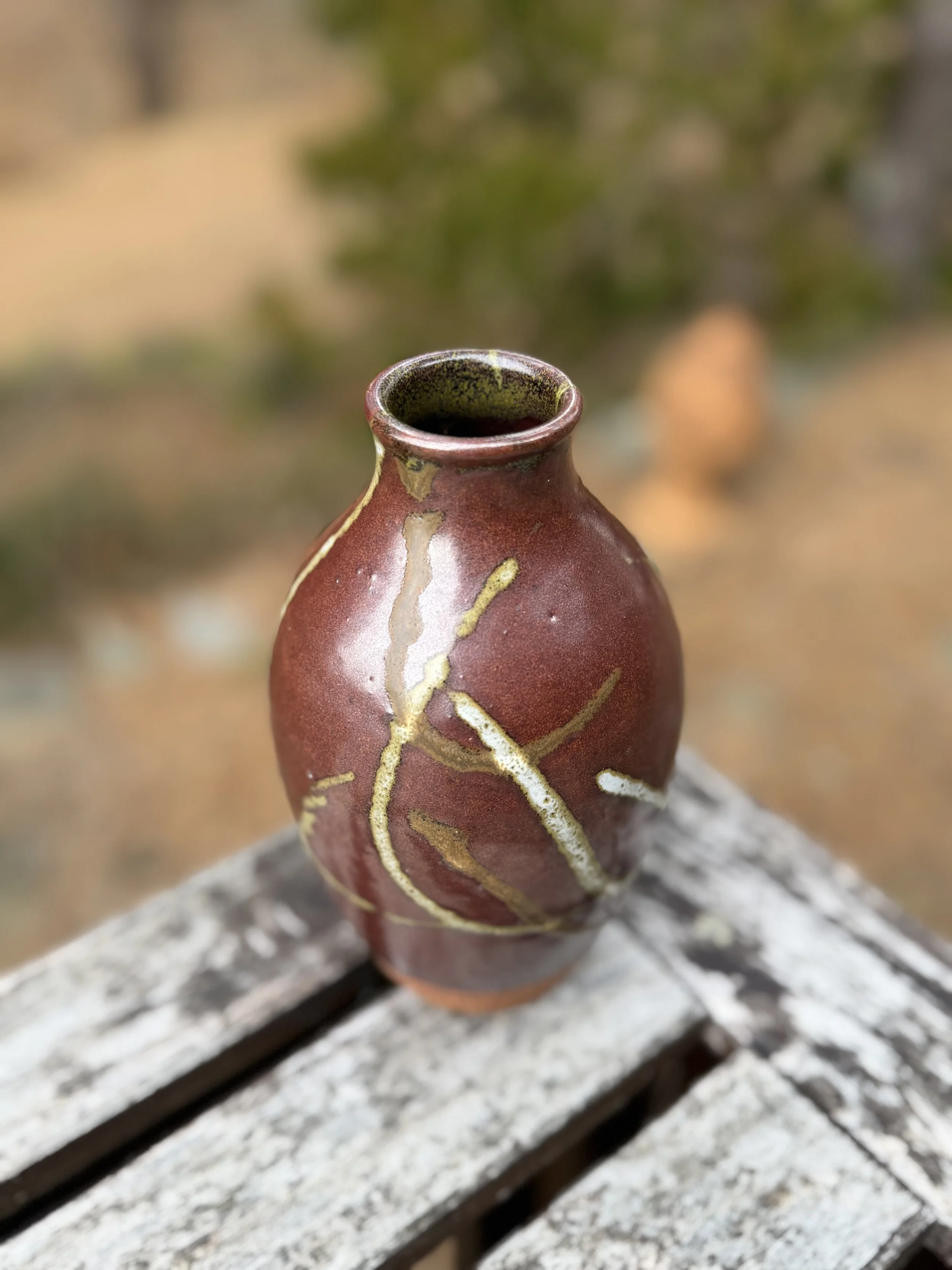 A small clay vase with a reddish-brown glaze and yellowish streaks, sitting on a weathered wooden surface outdoors.