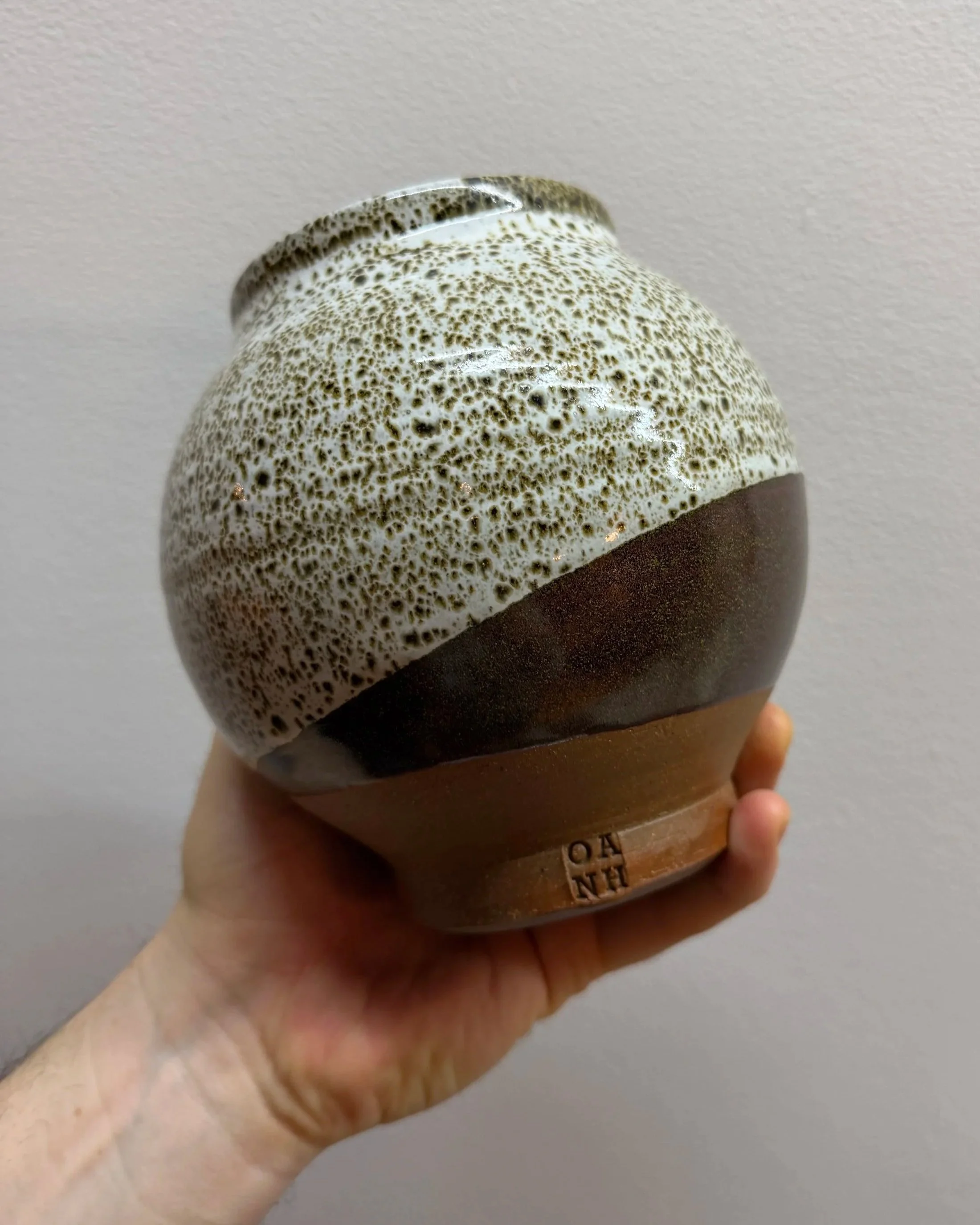 A hand holding a round, glazed ceramic mug with a speckled white and black upper part and a solid dark lower part, against a plain light-colored background.