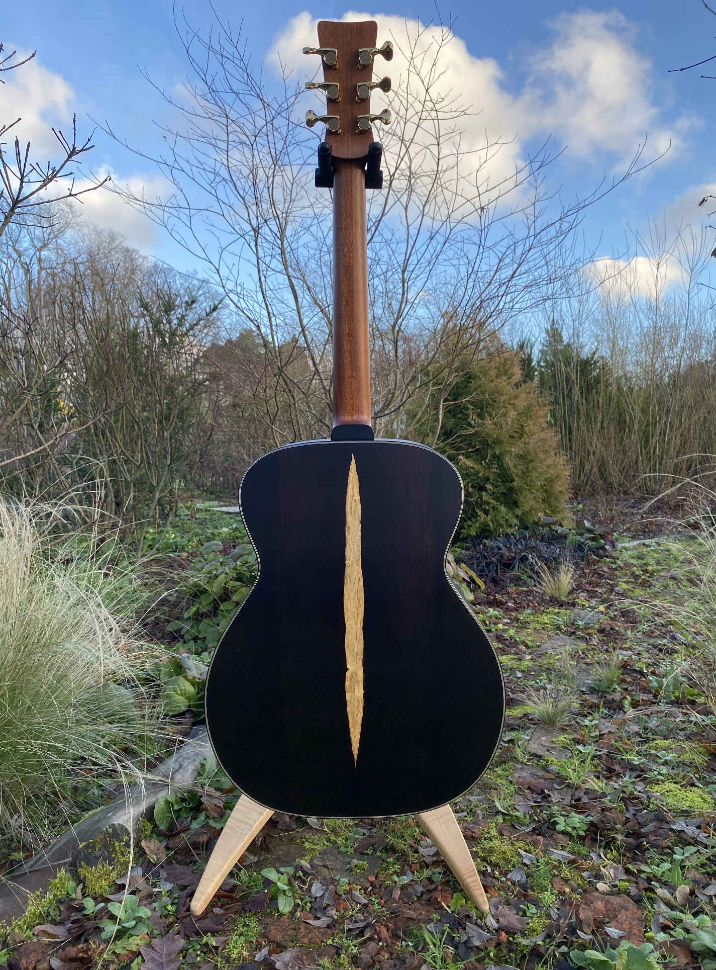 An acoustic guitar standing on a wooden stand outdoors in a garden with trees and bushes, under a partly cloudy sky.