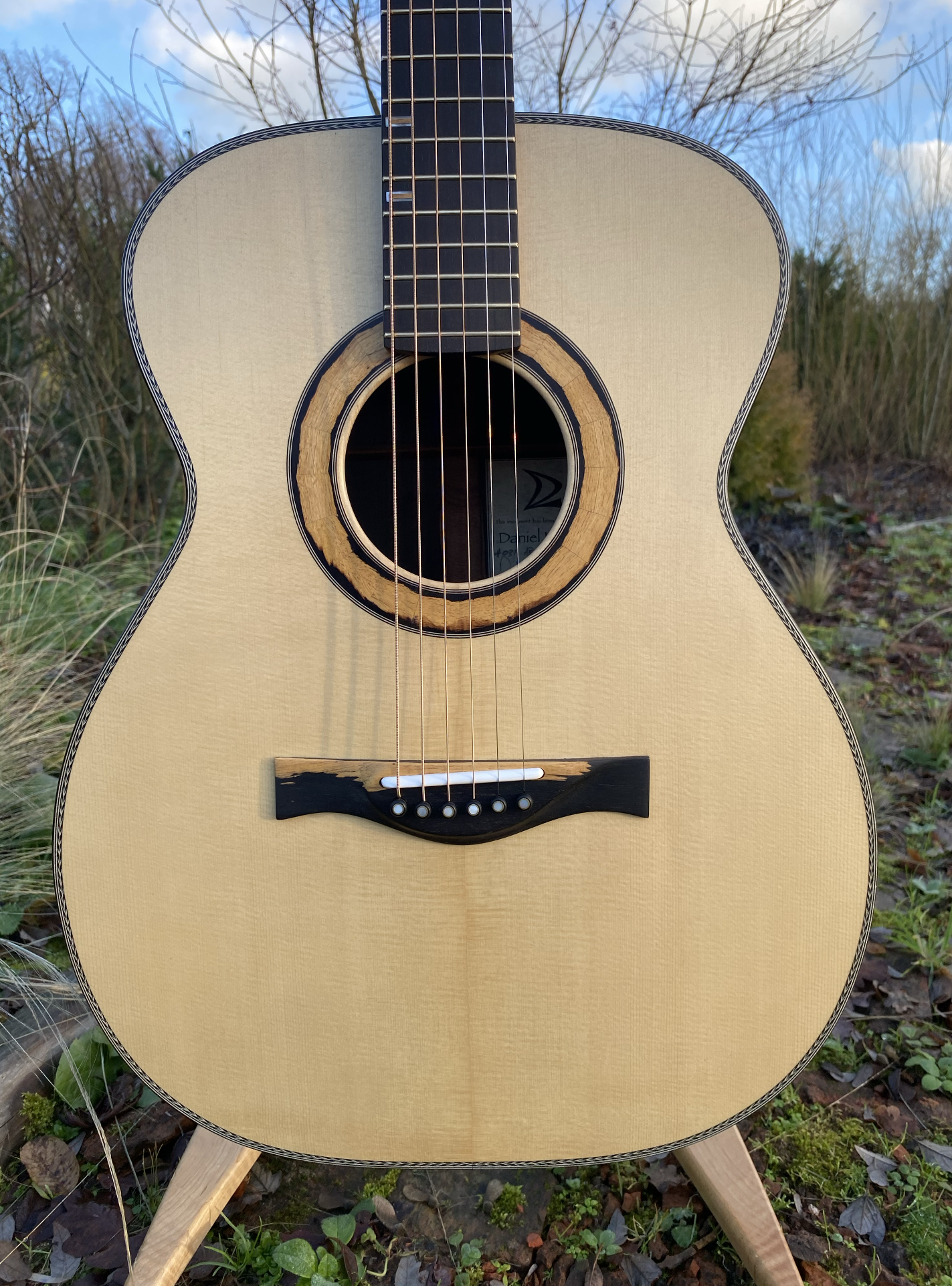 A close-up of an acoustic guitar outdoors on grass with trees and blue sky in the background, showing the body and soundhole.