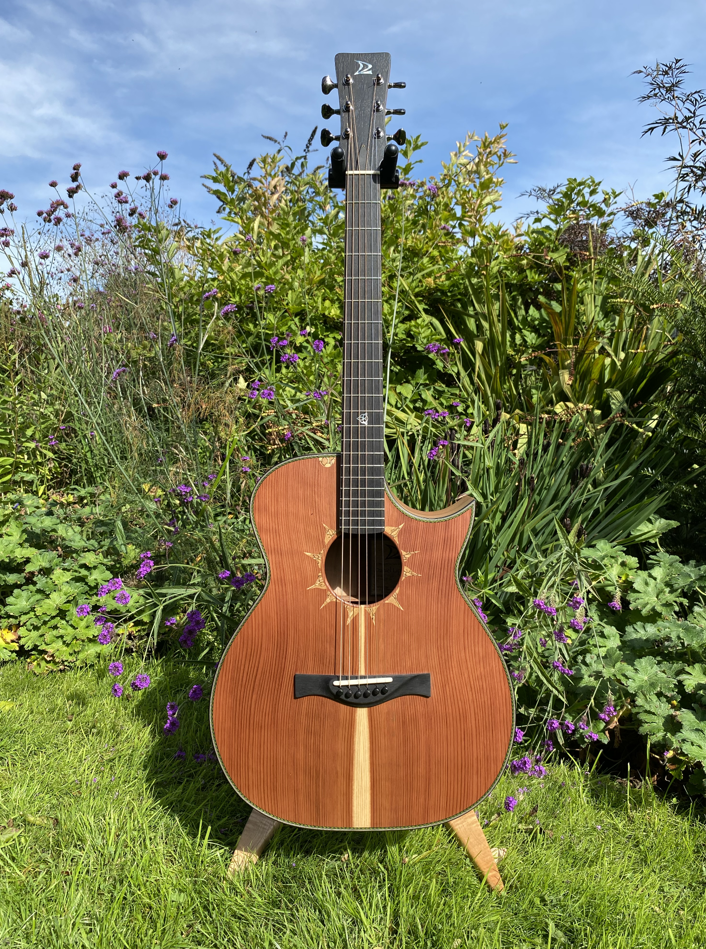 An acoustic guitar with a natural wood finish, black bridge and fretboard, standing on a garden lawn with purple flowers and green foliage, under a partly cloudy blue sky.