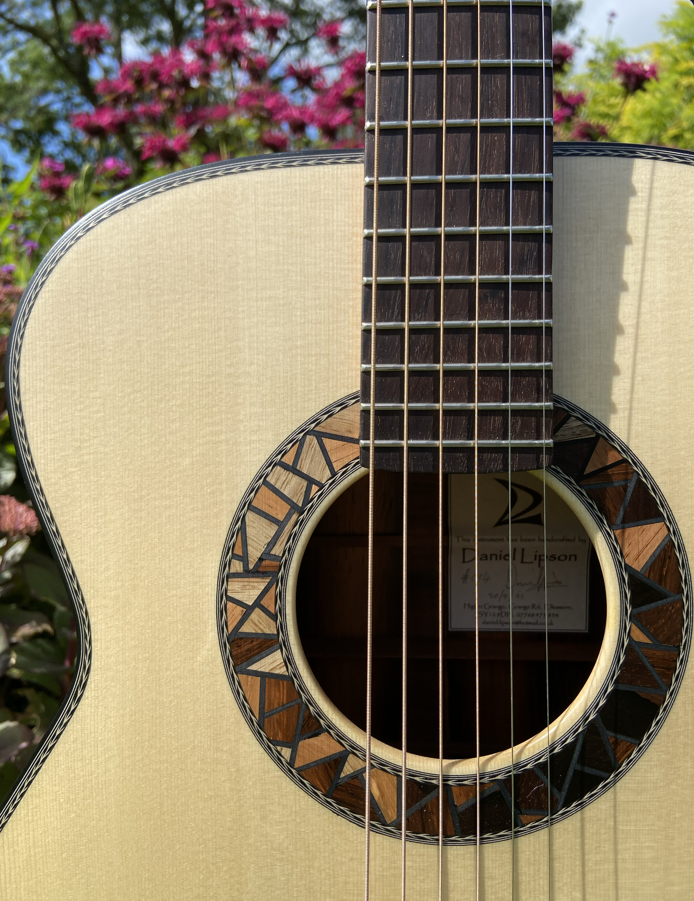 Close-up of an acoustic guitar with a beige body, dark fretboard, and decorative wood and inlay around the sound hole, with pink flowers and green foliage in the background.