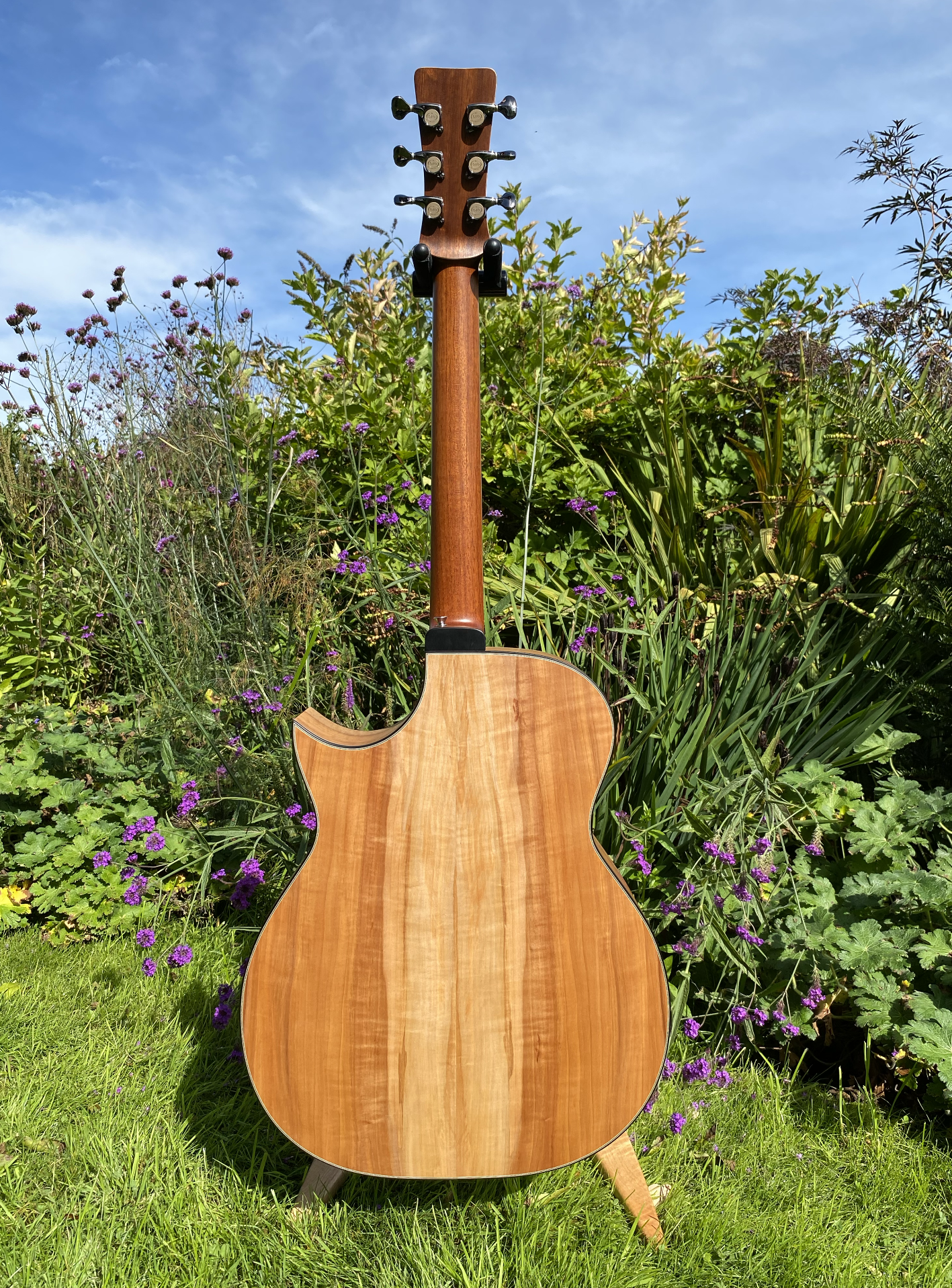 Back of an acoustic guitar standing on a grassy lawn with lush green plants and purple flowers in the background under a partly cloudy sky.
