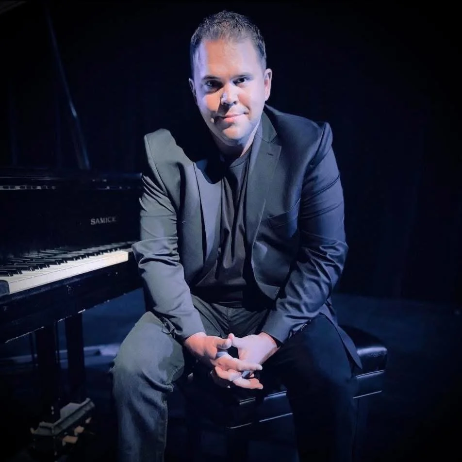 A man sitting on a piano bench beside a grand piano, wearing a dark suit and sitting with his hands clasped, in a dimly lit room.