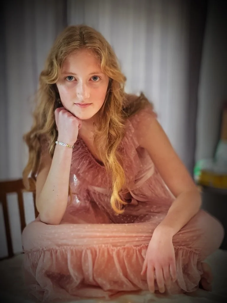 A young woman with long, wavy red hair sitting on a bed, resting her head on her hand and looking at the camera. She is wearing a pink, ruffled dress.
