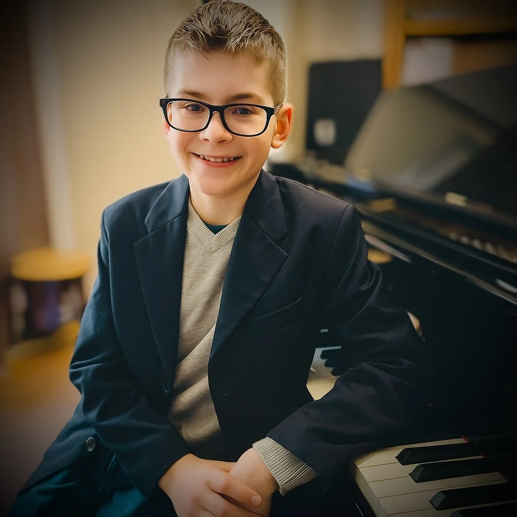 A smiling young boy with glasses wearing a blazer and sitting at a piano.