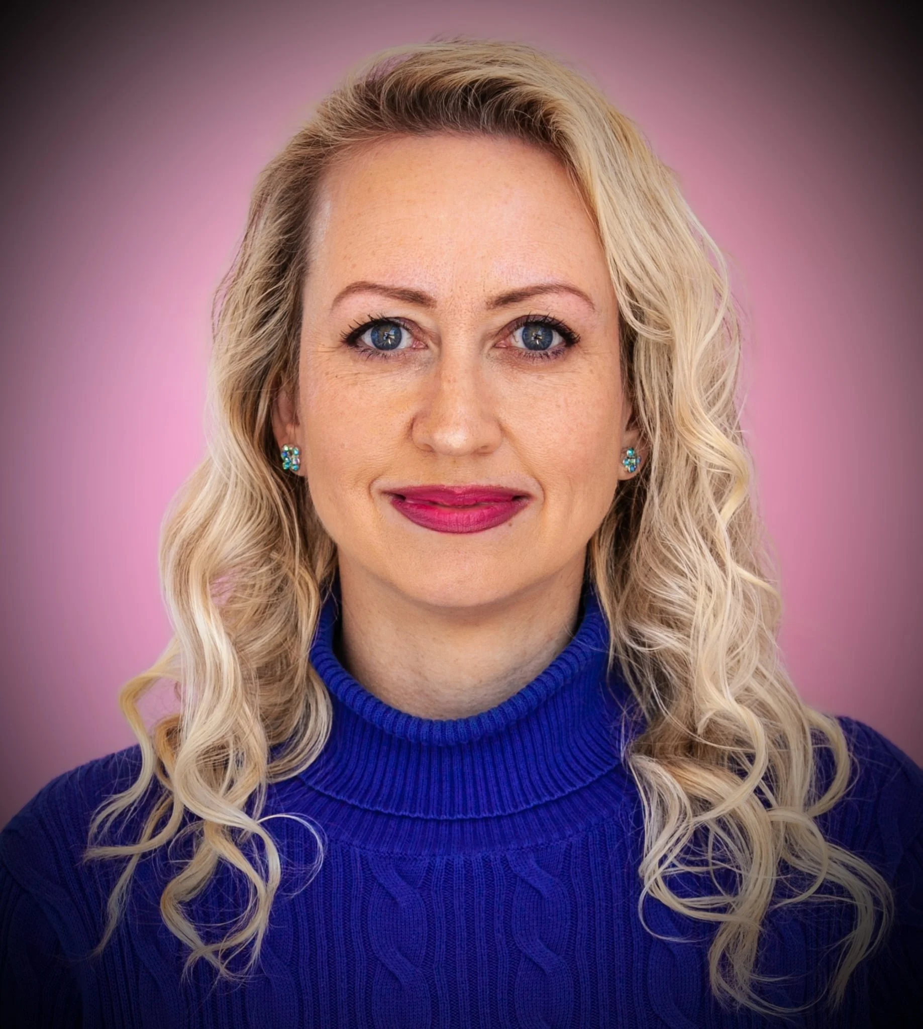 Close-up portrait of a woman with blond curly hair, blue eyes, wearing a blue turtleneck sweater, blue earrings, and pink lipstick, with a pink background.