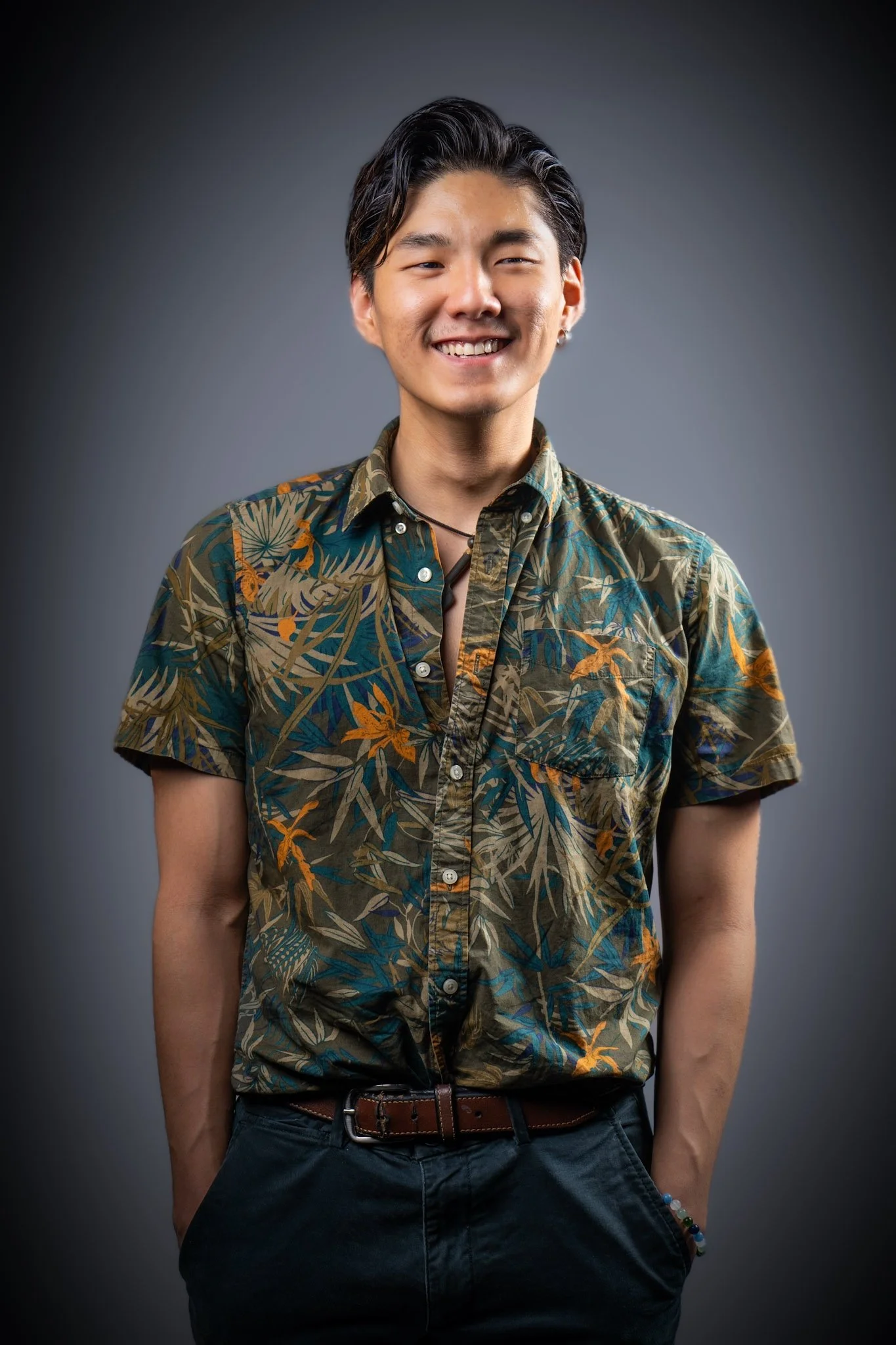 A young man with dark hair, smiling, wearing a tropical printed button-up shirt and dark pants, standing with hands in pockets against a plain dark background.