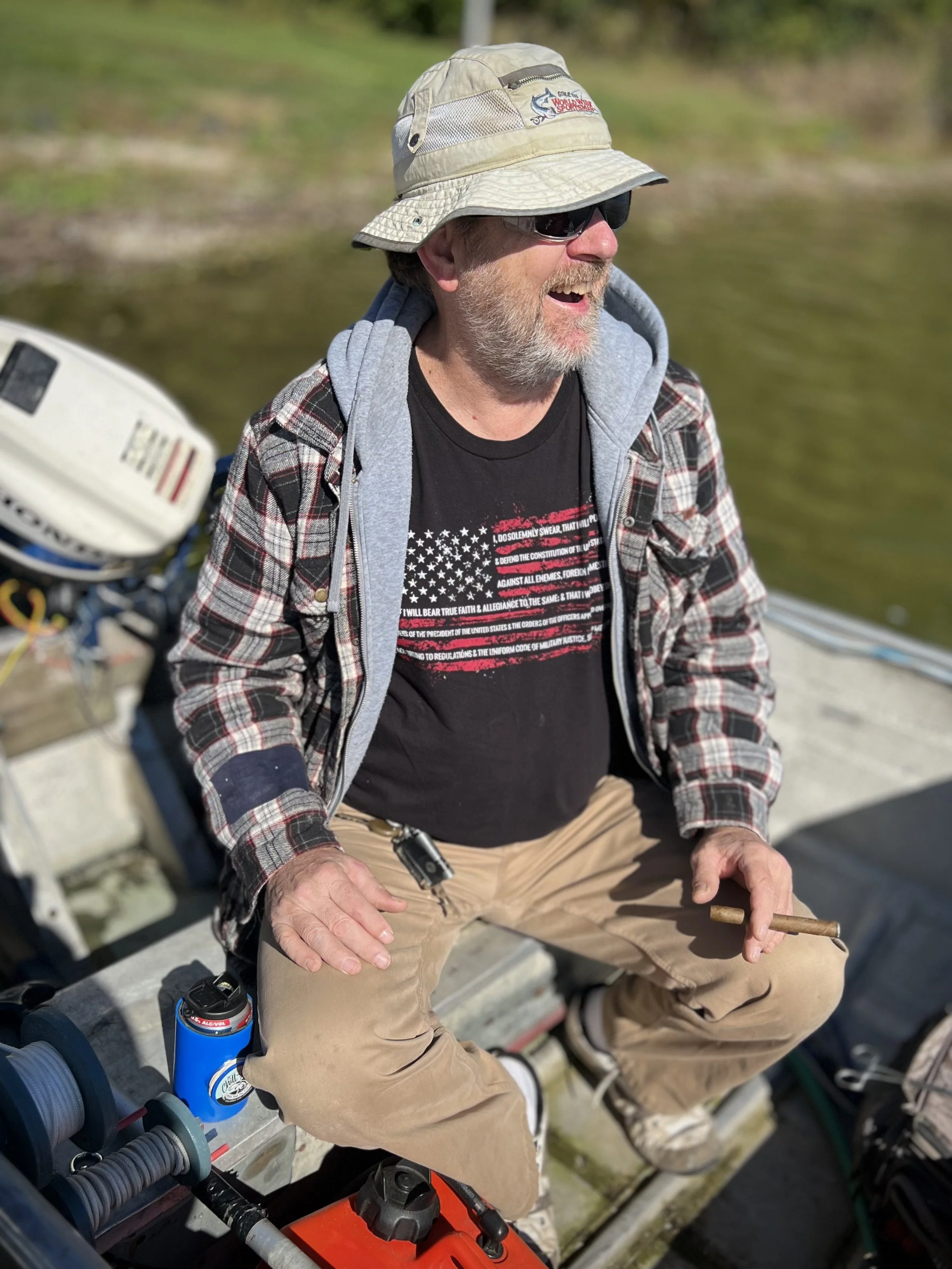 Man with a grey beard and sunglasses laughing while sitting outdoors on a boat, wearing a plaid jacket, a grey hoodie, a black t-shirt with an American flag graphic, a beige bucket hat, and holding a cigarette.