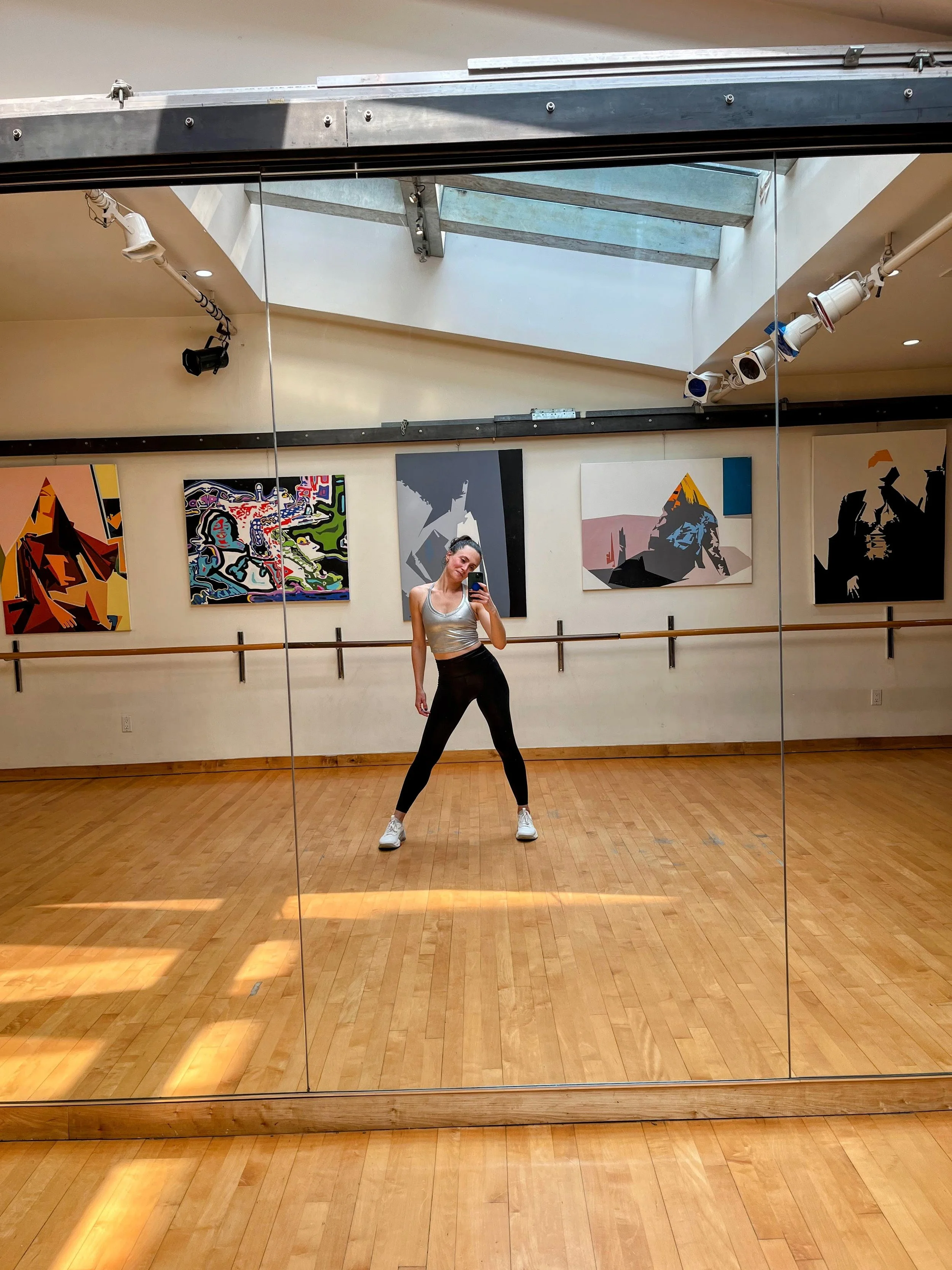 A woman in workout clothing taking a selfie in a dance studio with artwork on the walls and a wooden floor.