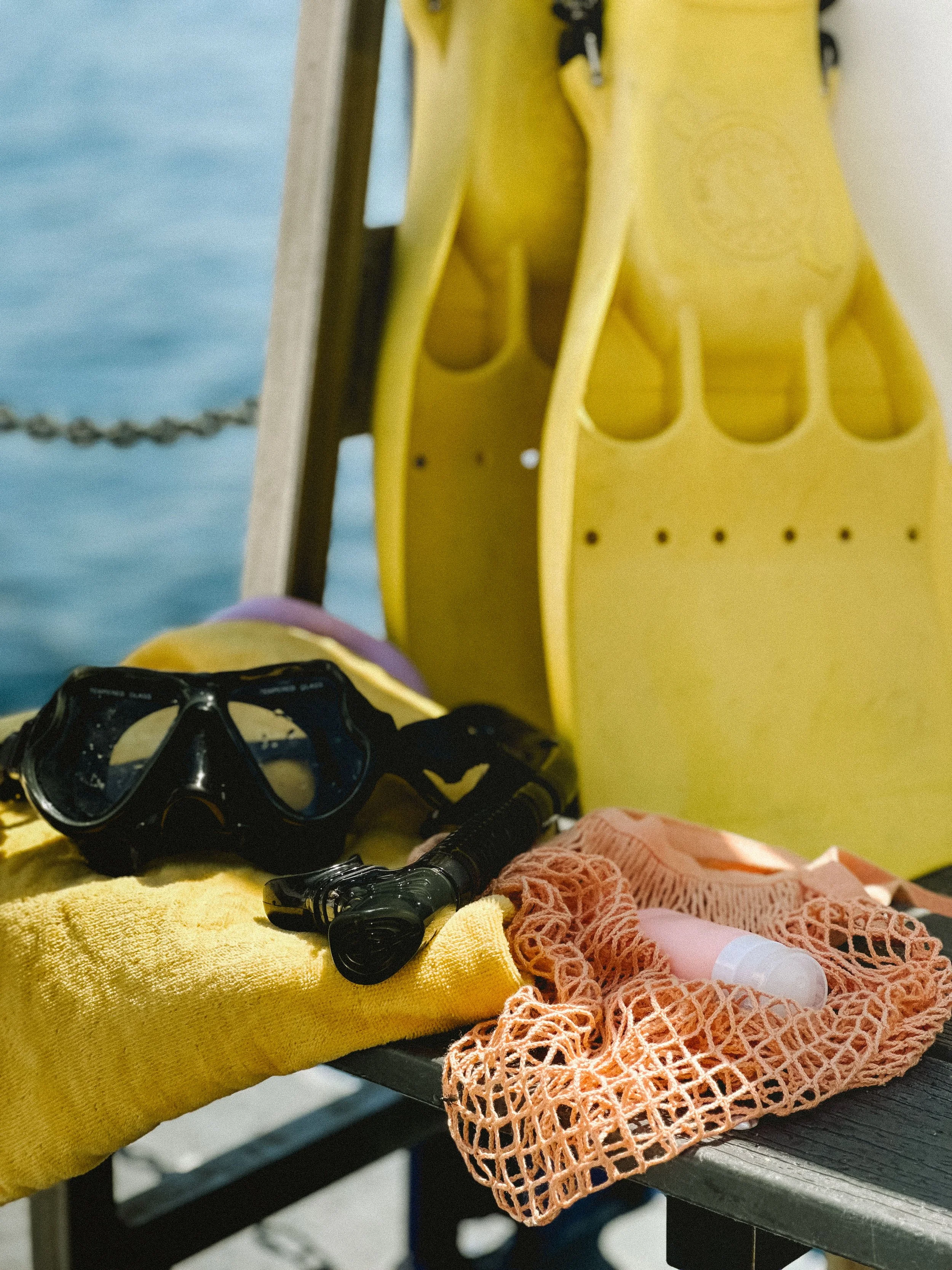 Scuba diving equipment including a mask, snorkel, and a small bottle, resting on a yellow towel with a yellow fin in the background near water.