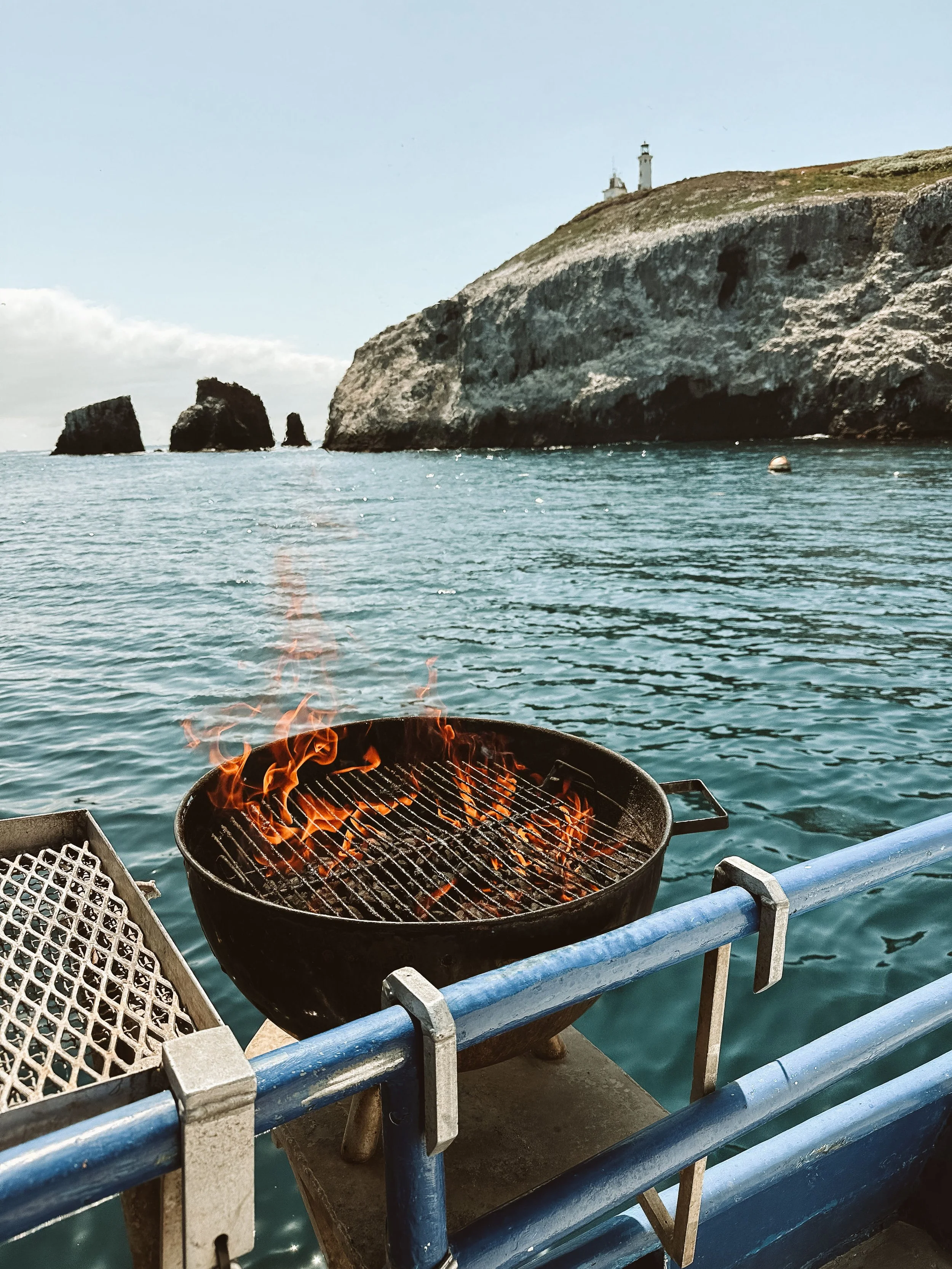Barbecue grill on a boat overlooking the ocean with rocky cliffs and lighthouses in the background.