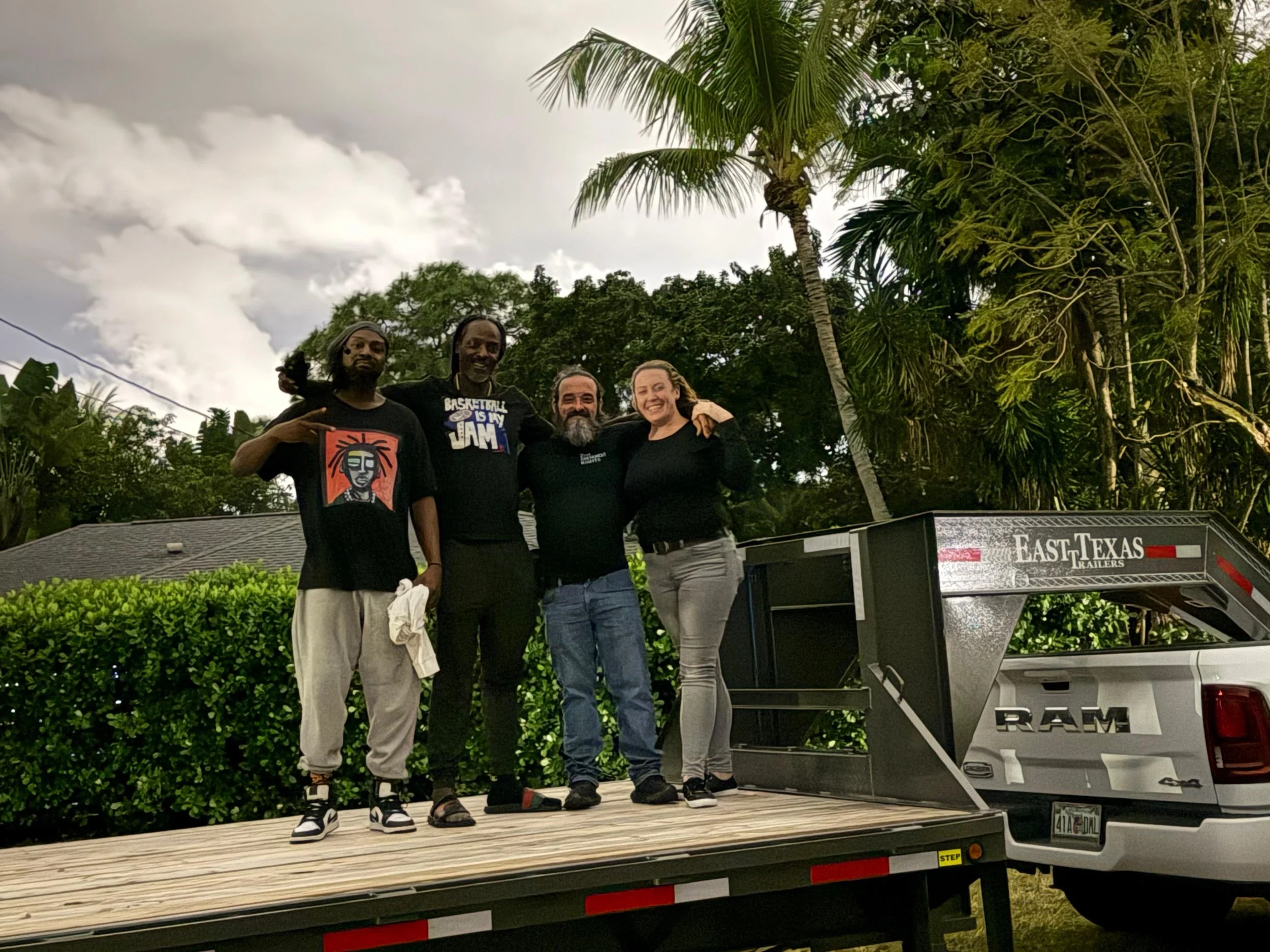 Four people standing on a wooden load bed in front of green bushes and palm trees, smiling and posing for the photo. One person is making a peace sign, and a trailer with the logo 'East Texas Trailers' is attached to a pickup truck.
