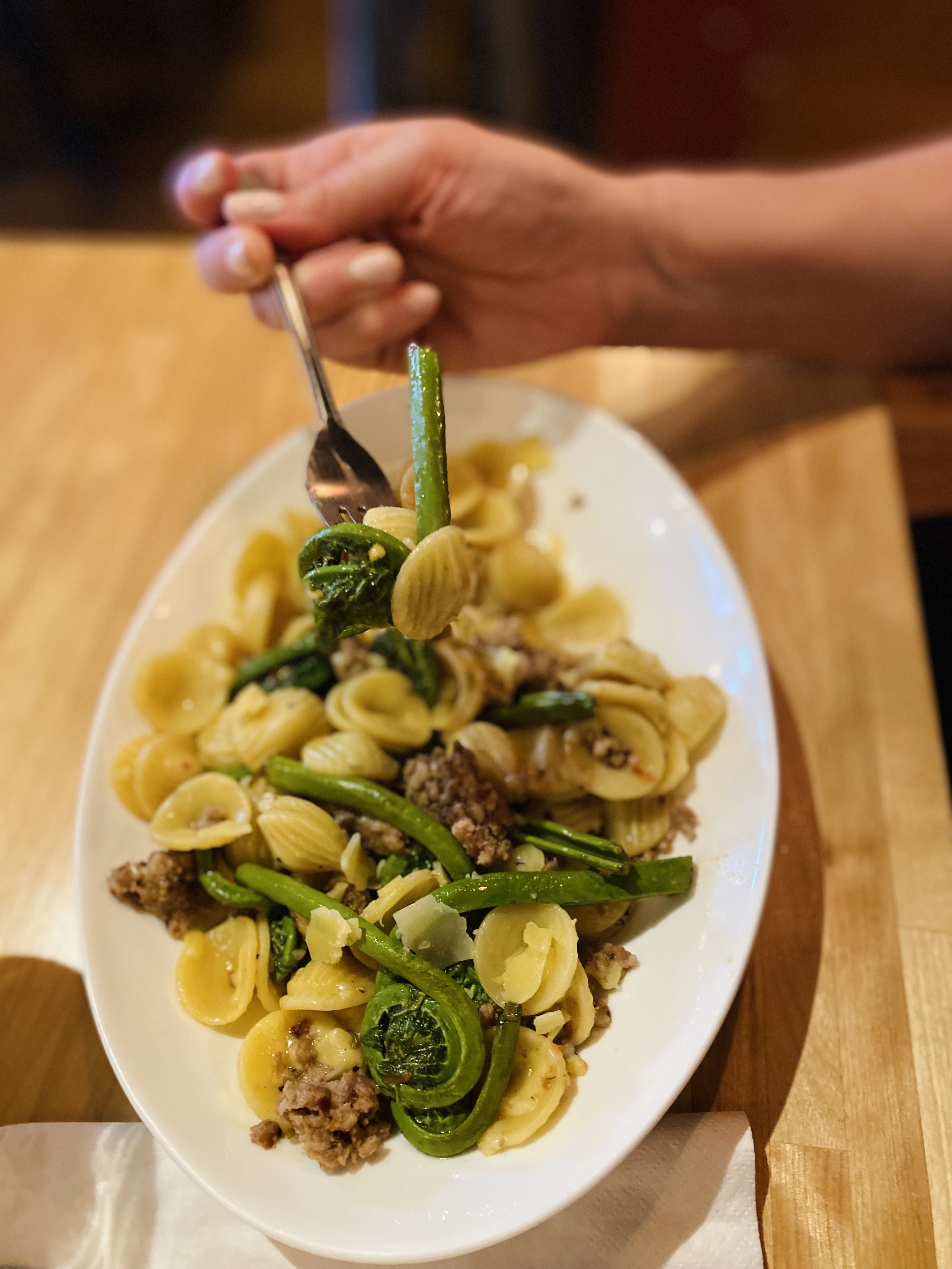 Person holding a fork with a bite of pasta on a plate of mixed pasta, green vegetables, and ground meat on a wooden table.