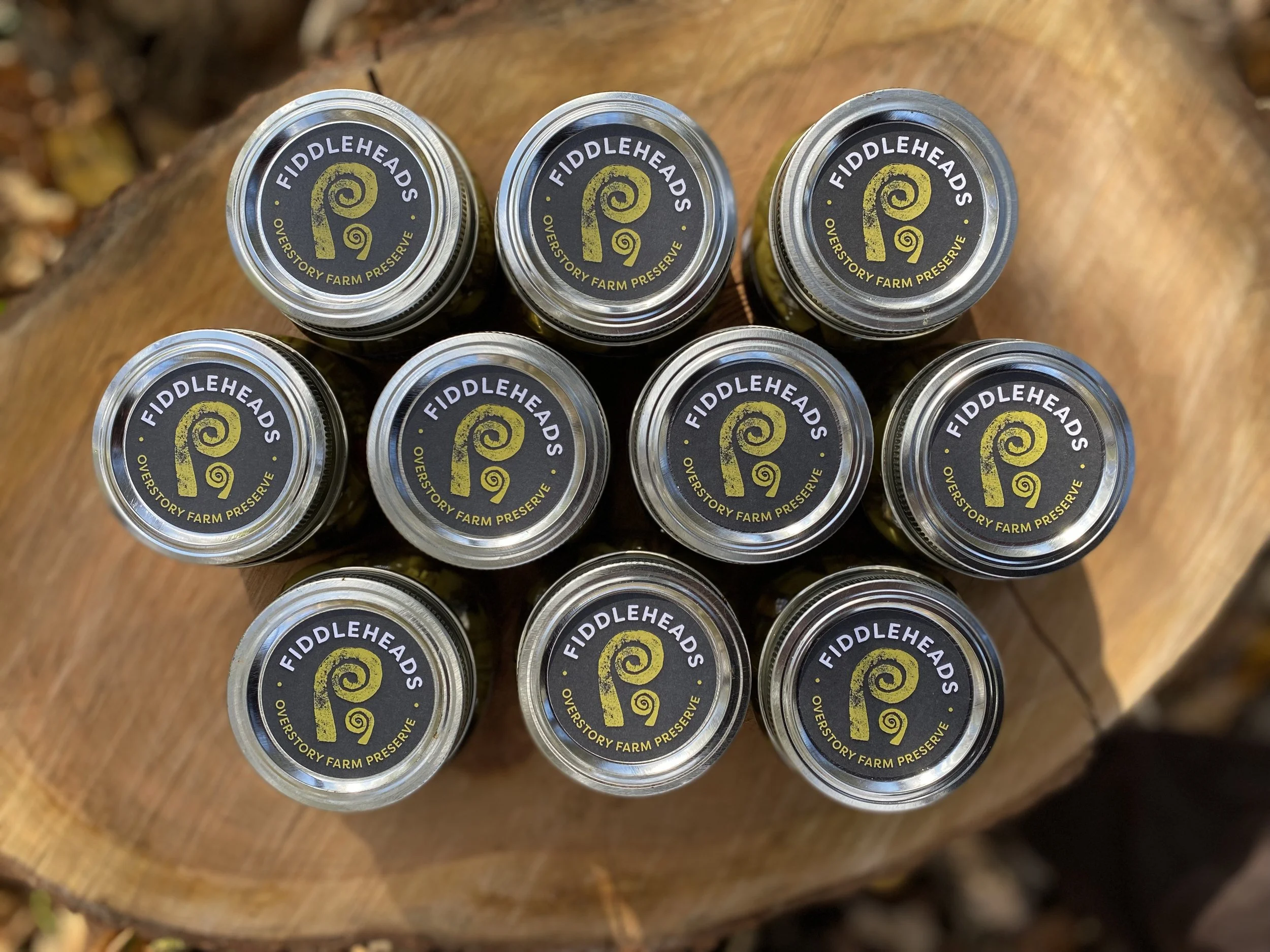 Top view of multiple jars with black and yellow labels reading 'Fiddleheads Overstory Farm Preserve' arranged on a wooden surface.