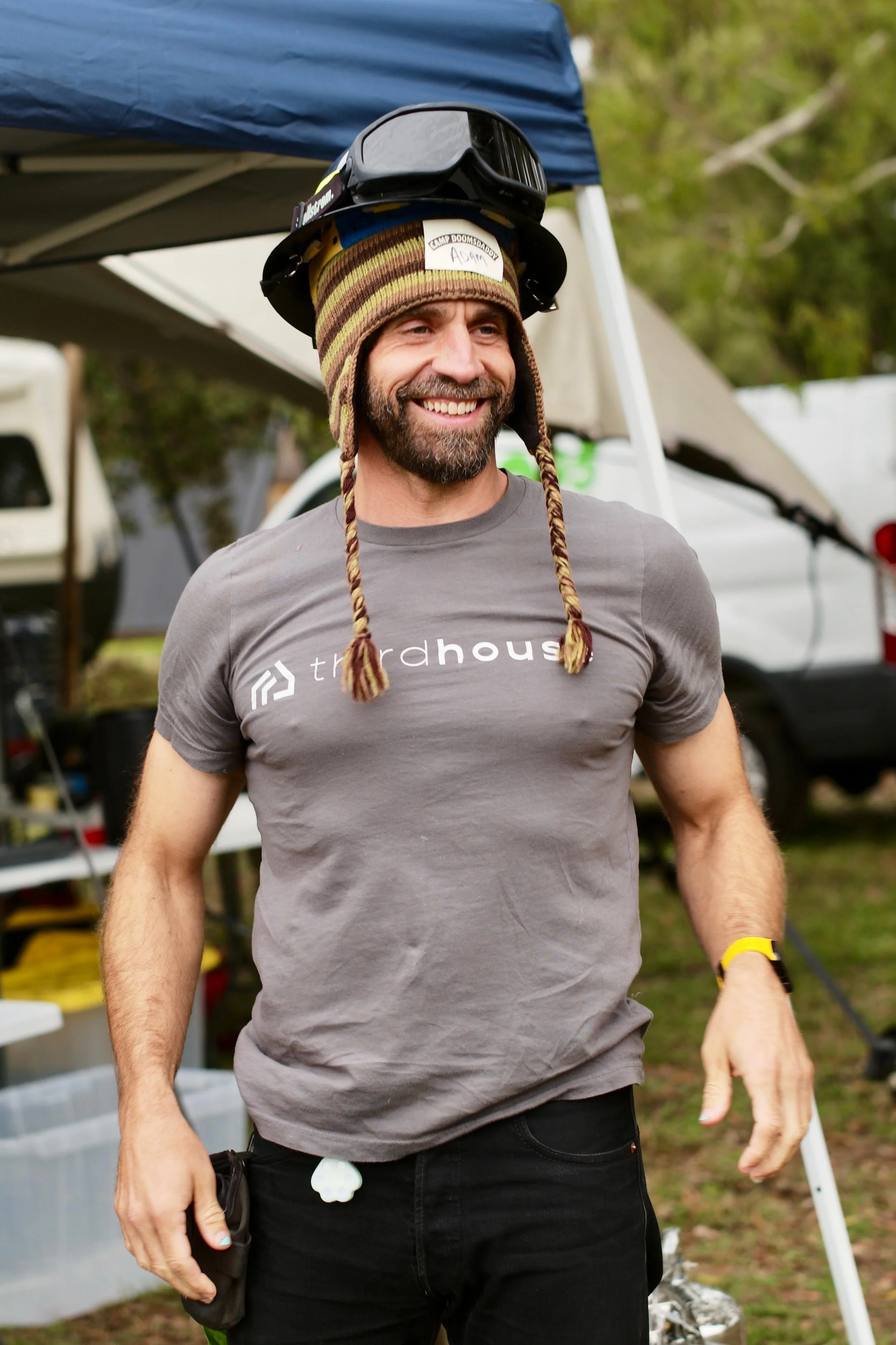 A smiling man wearing a gray t-shirt with the word 'thirdhouse' printed on it, a colorful knit beanie with earflaps, and a pair of sunglasses on his head, standing outdoors near tents and vehicles.