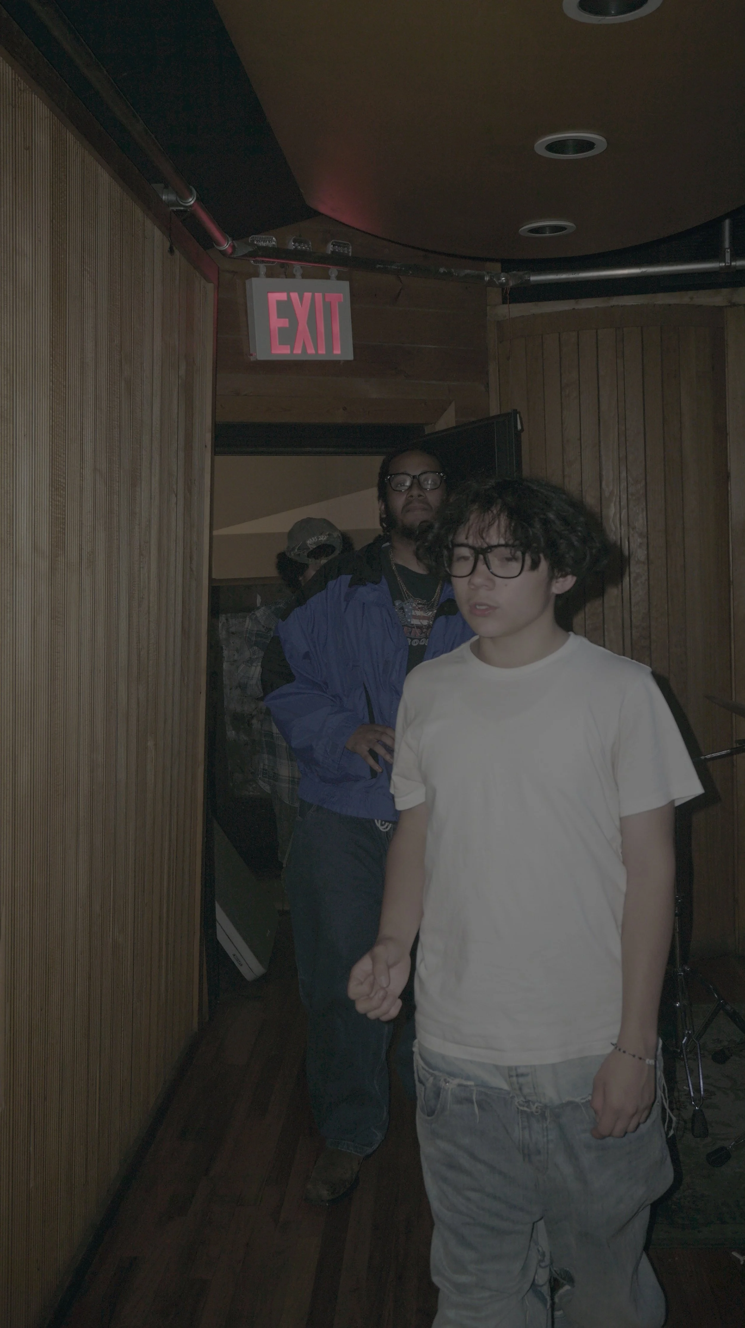 Two young men with curly hair and glasses standing in a dimly lit room with wood-paneled walls, one in a white T-shirt and the other in a blue jacket, near an exit sign.